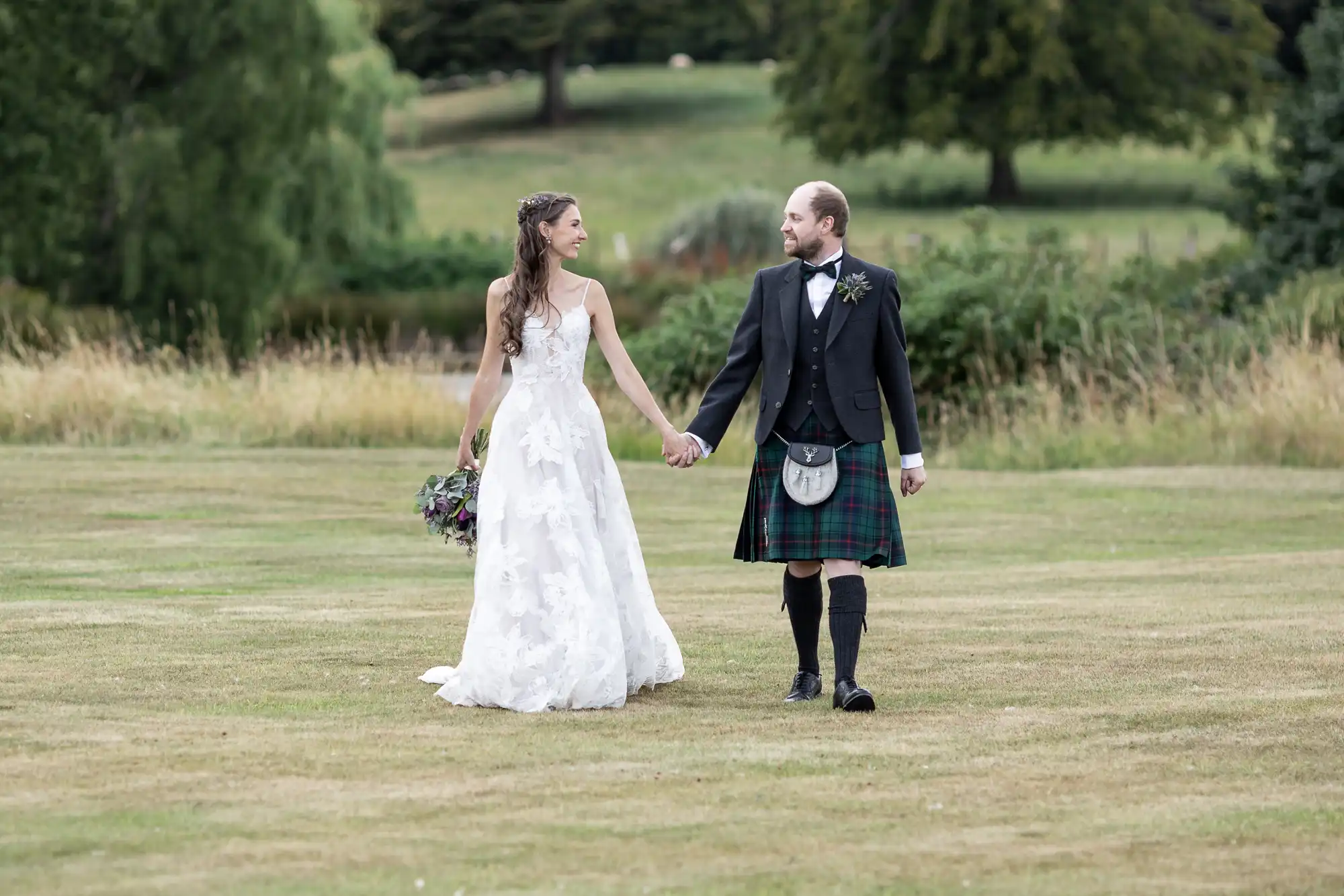 A bride in a white dress and groom in a kilt walk hand-in-hand on a grassy field.