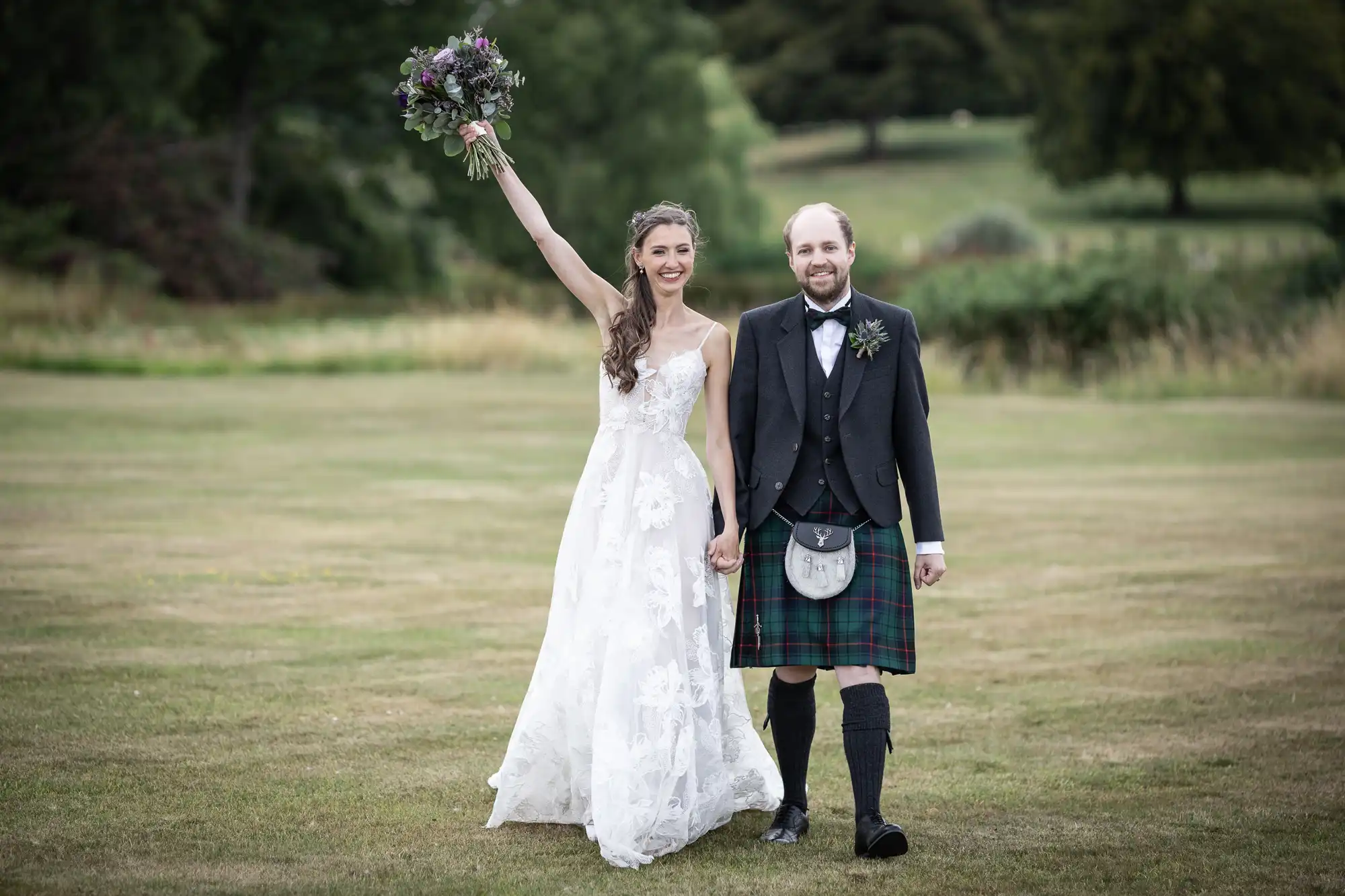 A bride in a white gown raising a bouquet walks hand-in-hand with a groom in a kilt and suit on green grass.