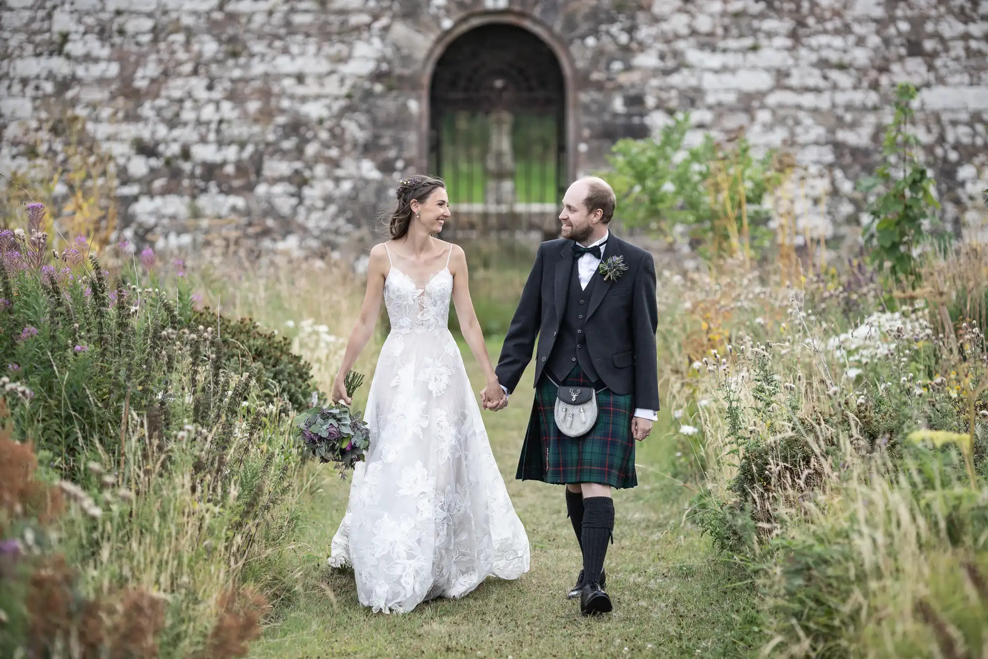 A bride in a white dress and a groom in a kilt walk hand-in-hand on a grassy path with an old stone building in the background.