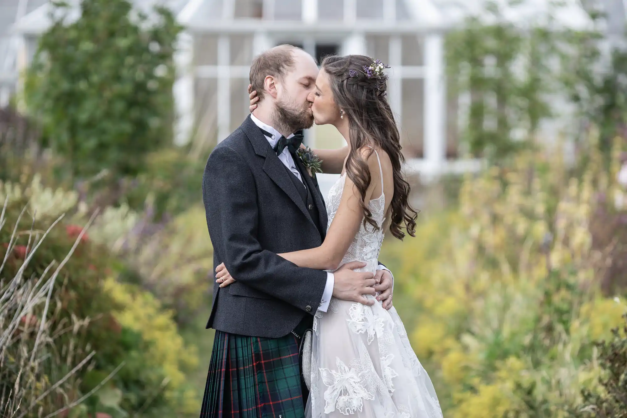 A bride and groom share a kiss outdoors, with the groom wearing a kilt and the bride in a white dress. A greenhouse and greenery are in the background.