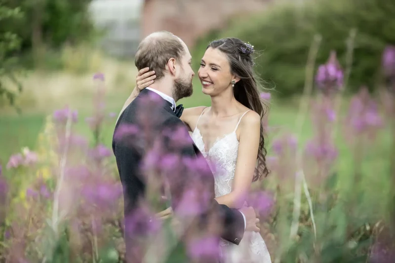 Summer wedding at Dunglass Estate: A couple dressed in wedding attire embrace in a garden, surrounded by blooming flowers. The woman is smiling while looking at the man.