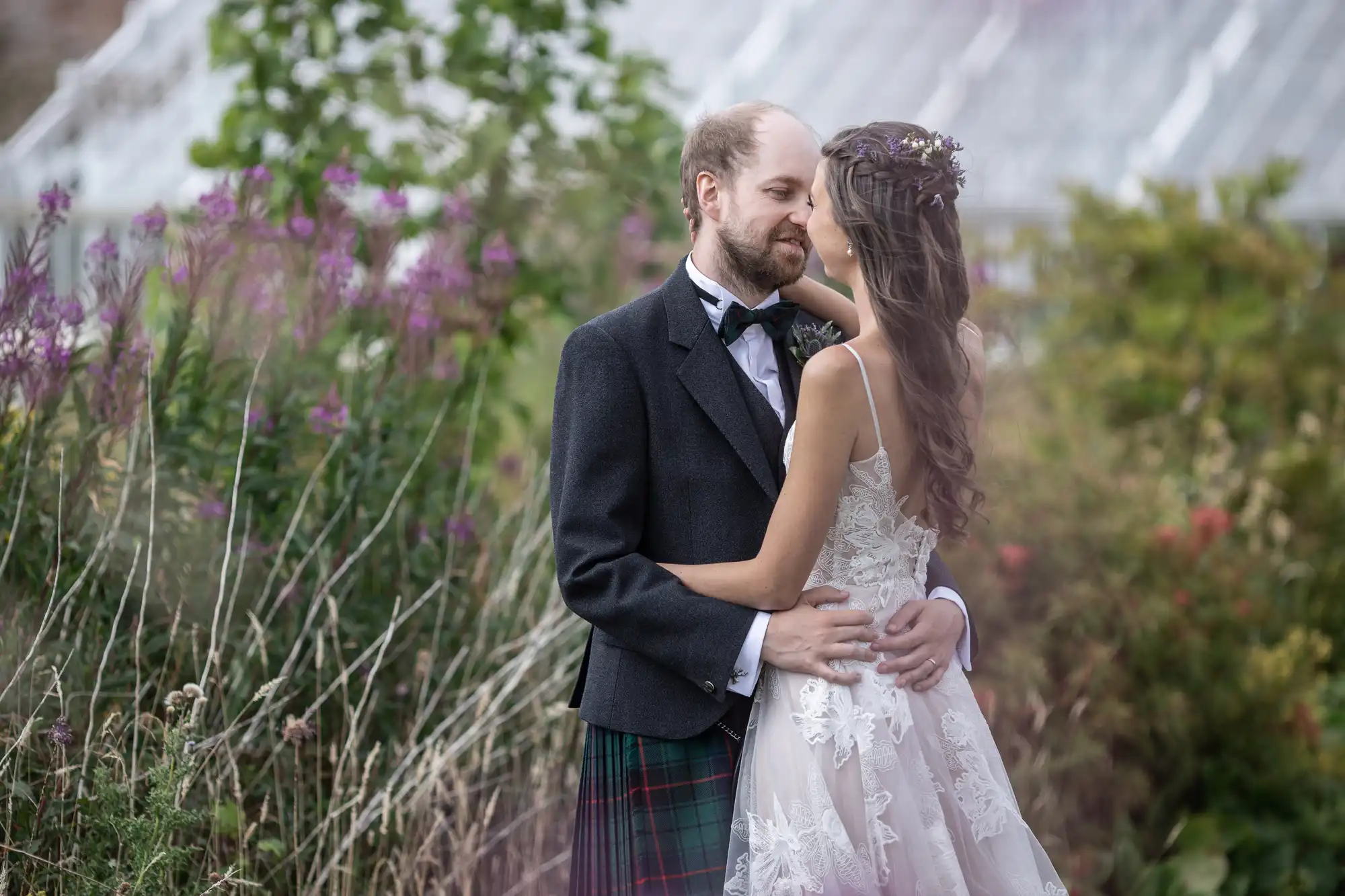 A couple embraces outdoors, the man in a suit with a kilt and the woman in a white wedding dress, surrounded by greenery and flowers.