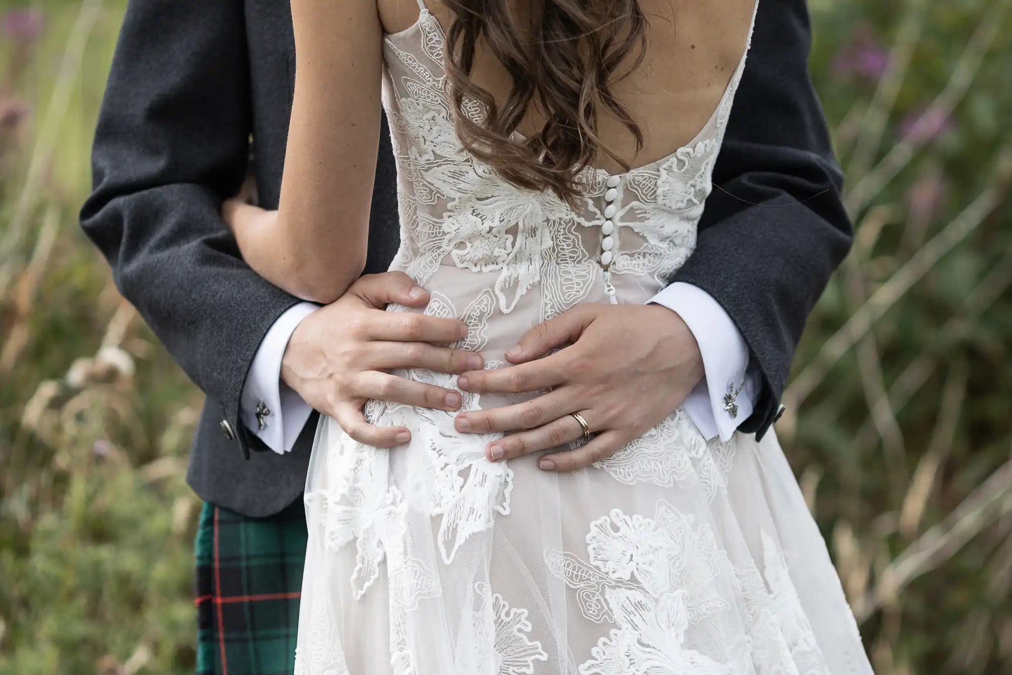 Close-up of a couple embracing with the woman's back facing the camera, wearing a white lace dress, while the man is dressed in a dark jacket and plaid trousers.