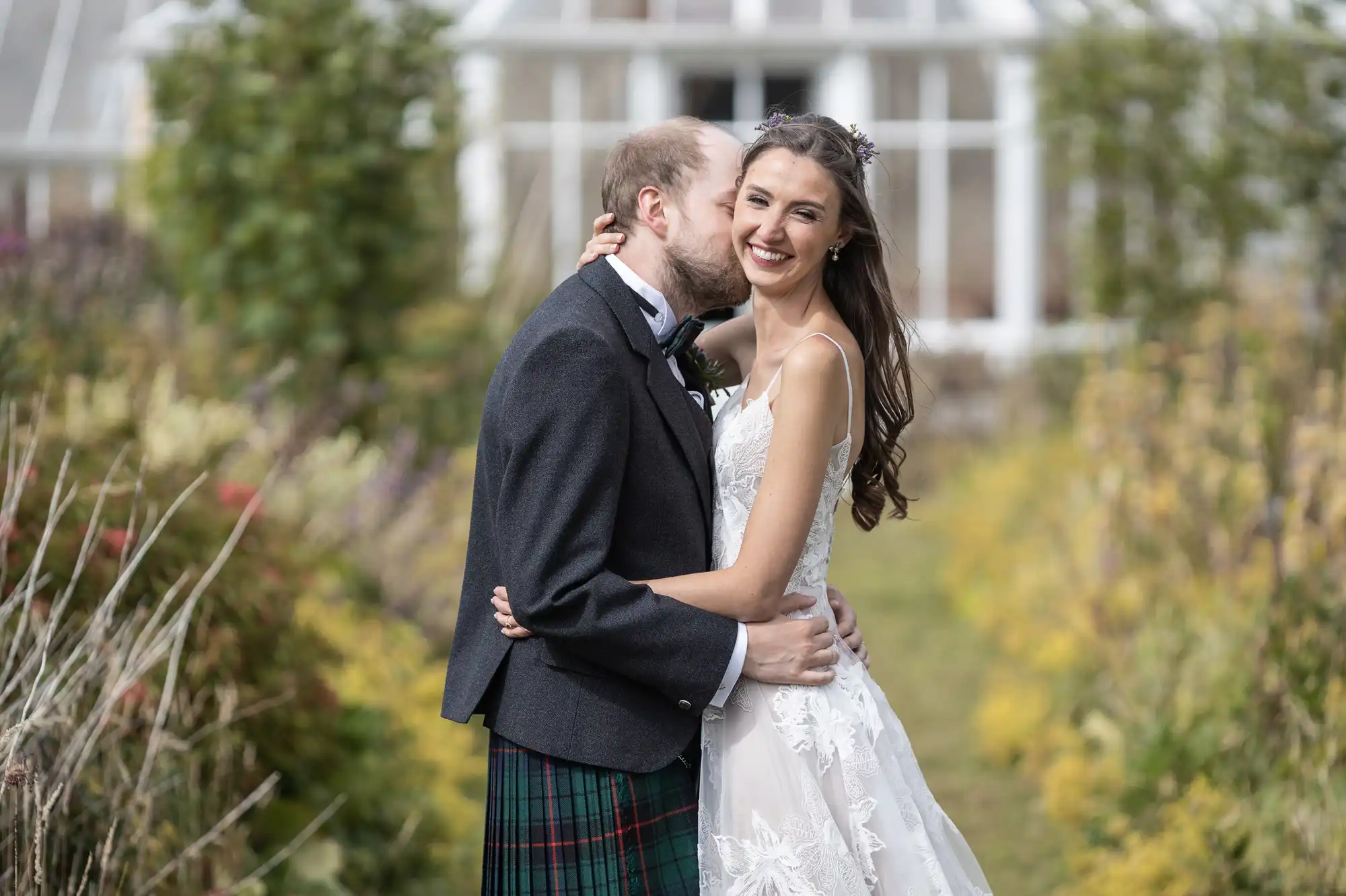 A man in a kilt and a woman in a white wedding dress share a joyful moment outdoors in a garden setting. The man leans in to kiss the woman's cheek, and both are smiling.