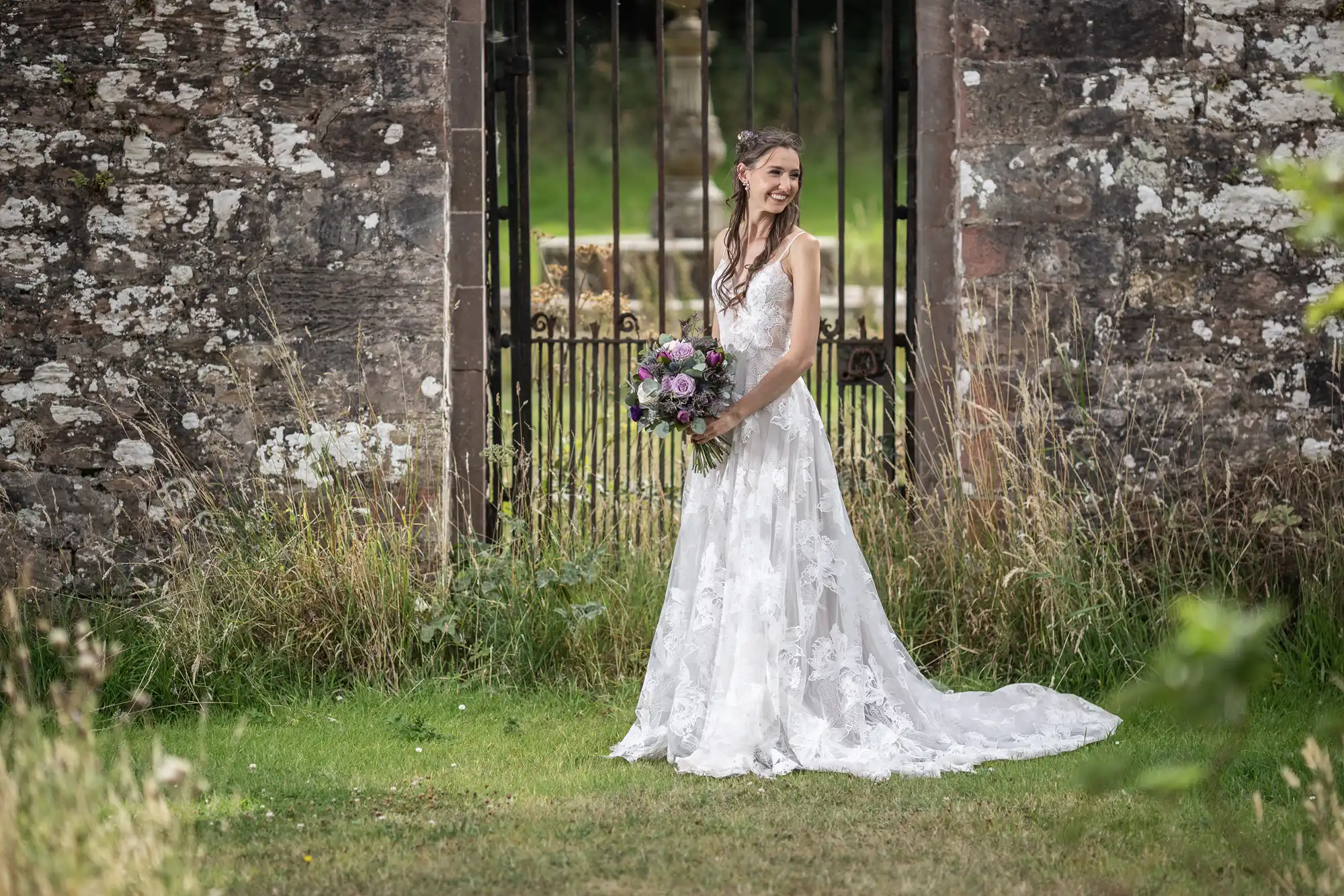 A bride in a white lace wedding gown holds a bouquet of flowers while standing on grass in front of a stone gate.