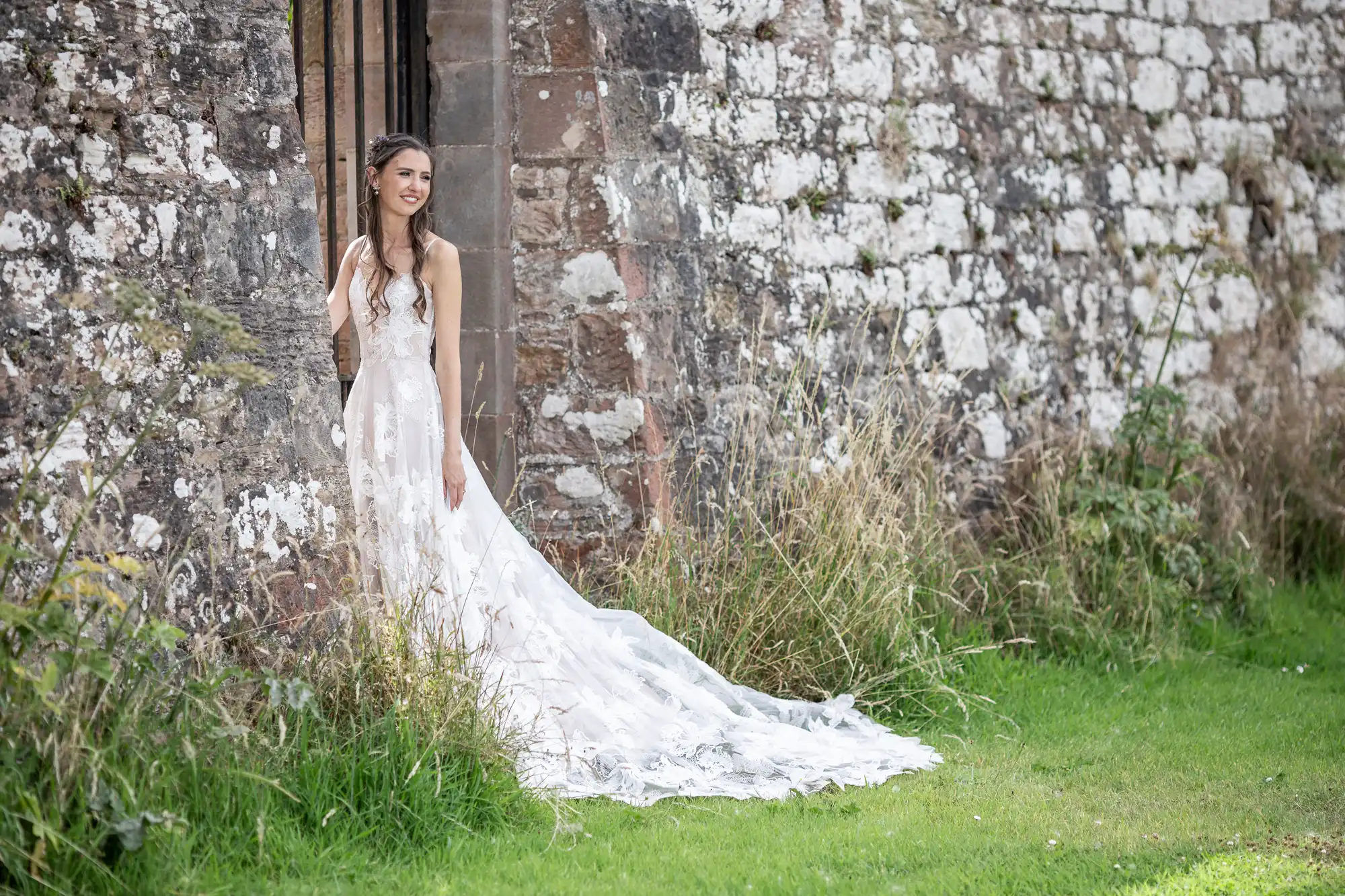 A woman in a long white wedding dress stands outdoors beside an old stone wall, looking to the side. She is holding onto the wall and standing on green grass surrounded by plants.