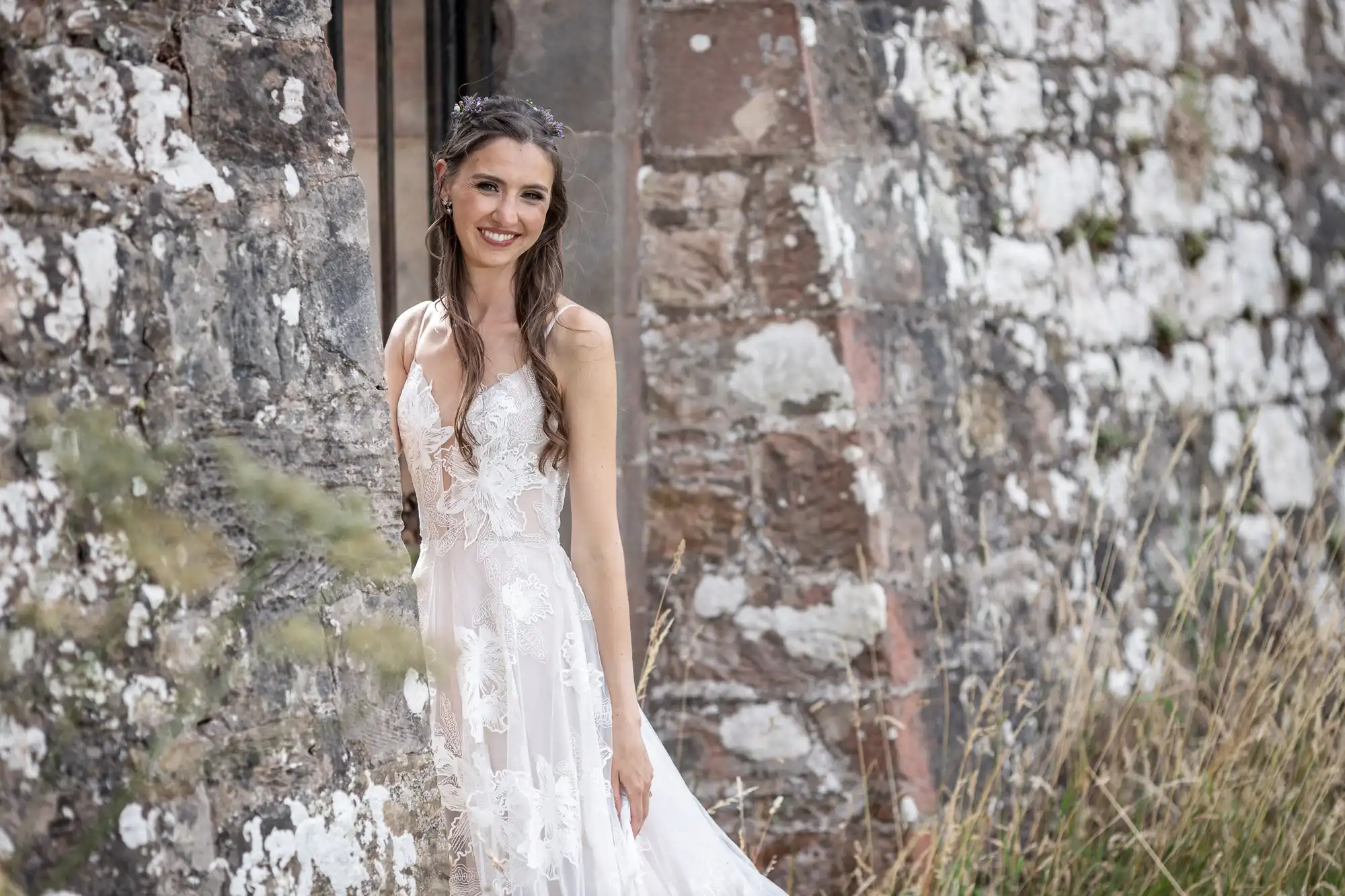 A woman in a white lace dress stands smiling against a weathered stone wall with greenery in the foreground.