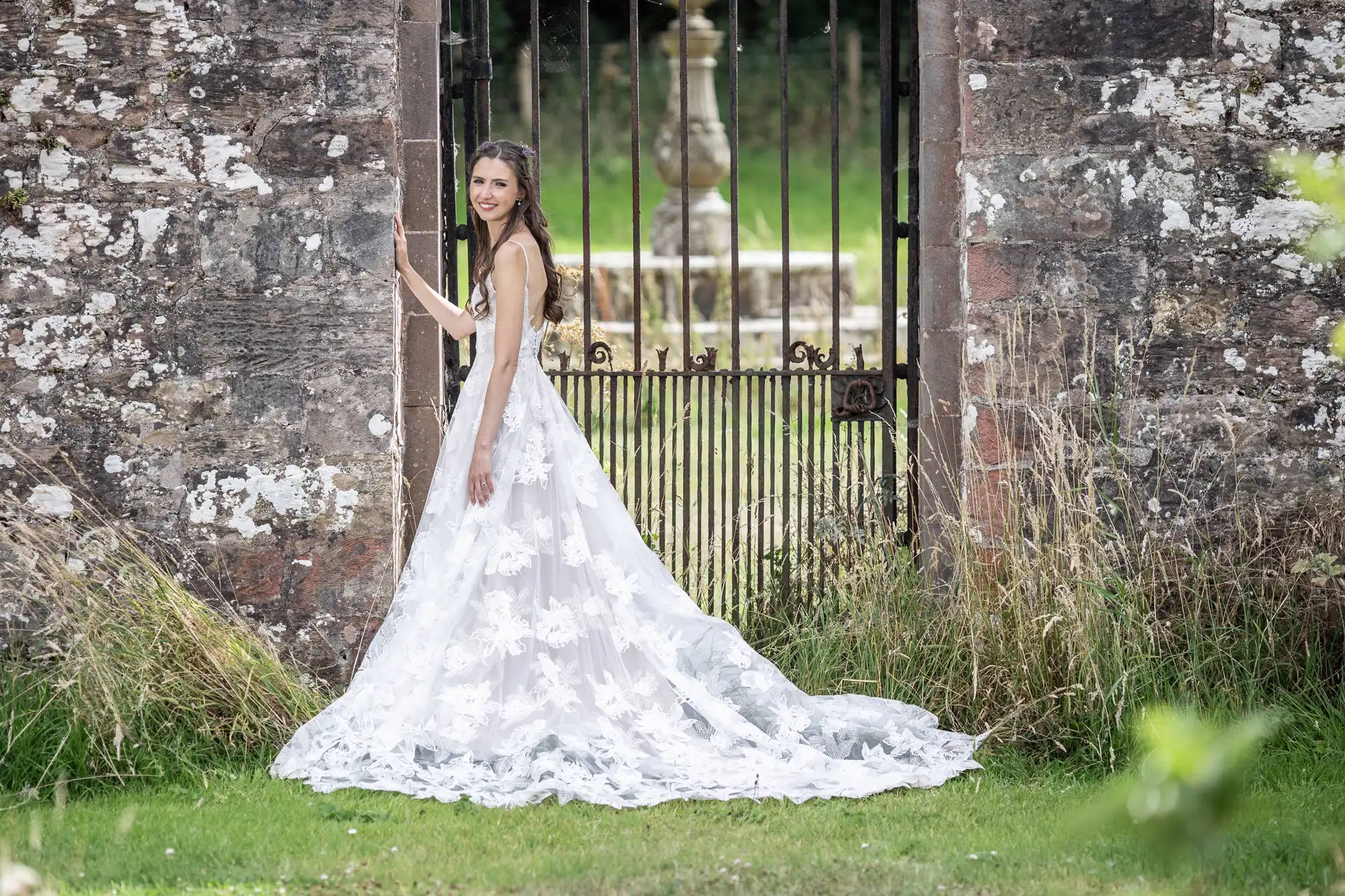 A woman in a white lace wedding dress stands next to a tall, old stone gate, with green grass and a blurred stone fountain visible in the background.