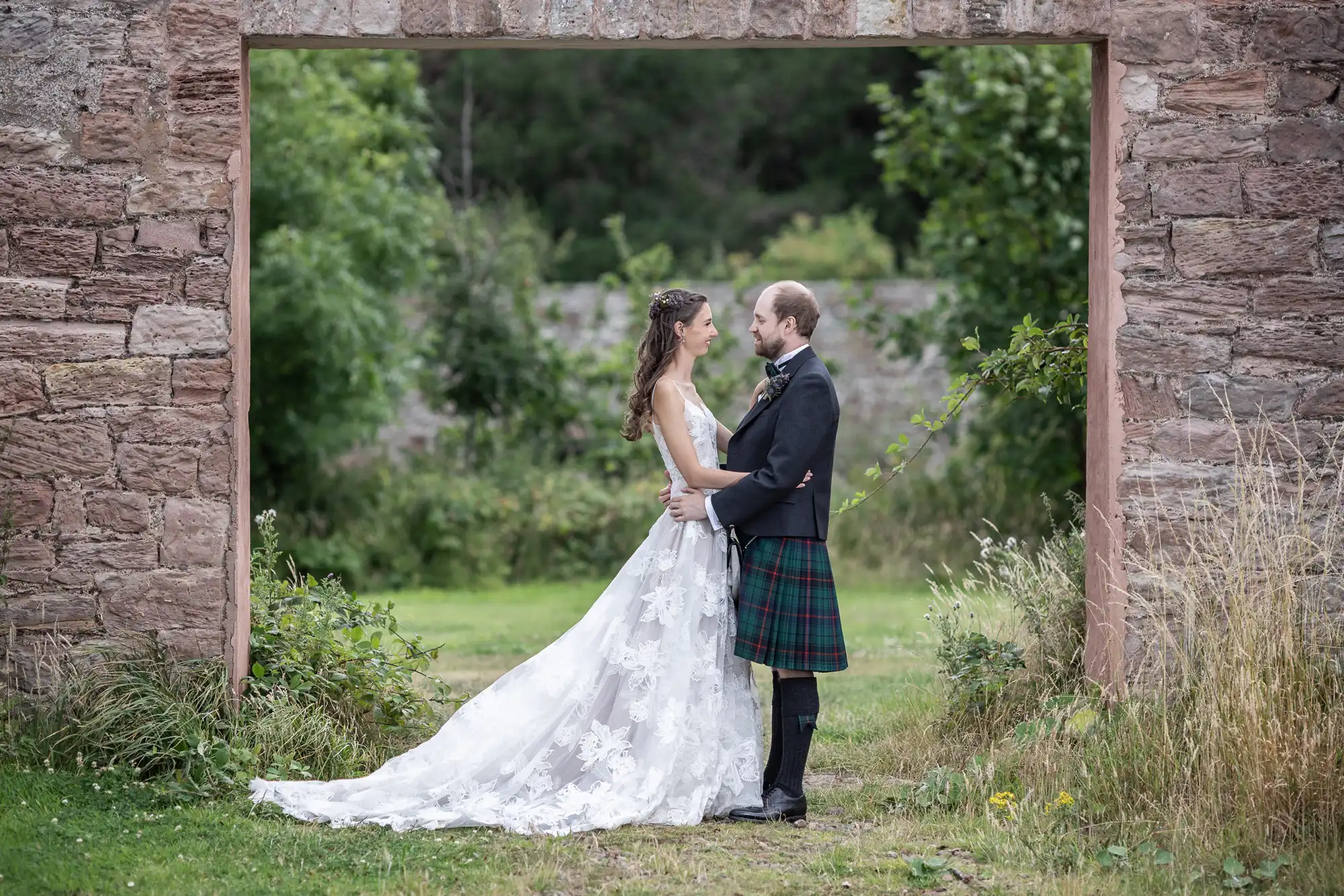 A couple, dressed in a wedding gown and a kilt, stands facing each other, holding hands in front of a stone archway with greenery in the background.