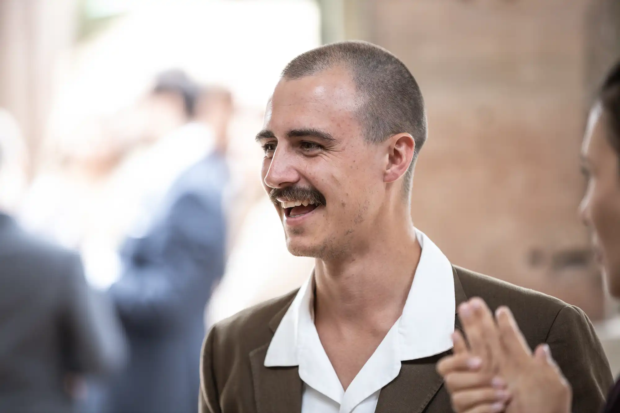 A man with a mustache, wearing a white-collared shirt and brown jacket, smiling and engaged in conversation at a social event.