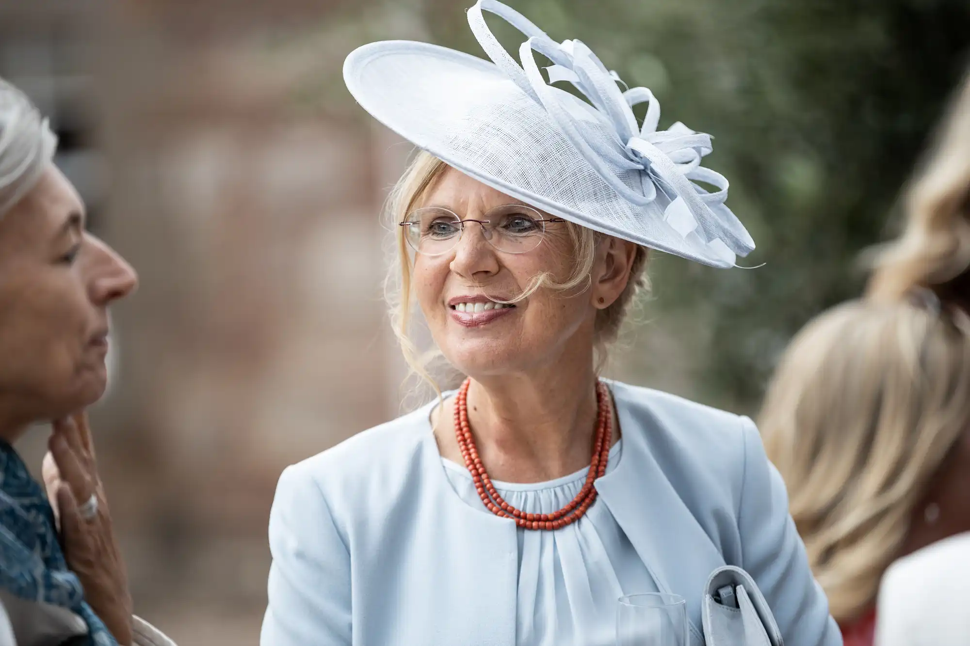 A woman with glasses, wearing a white hat and light blue outfit, stands outdoors engaged in conversation, with blurred figures and greenery in the background.