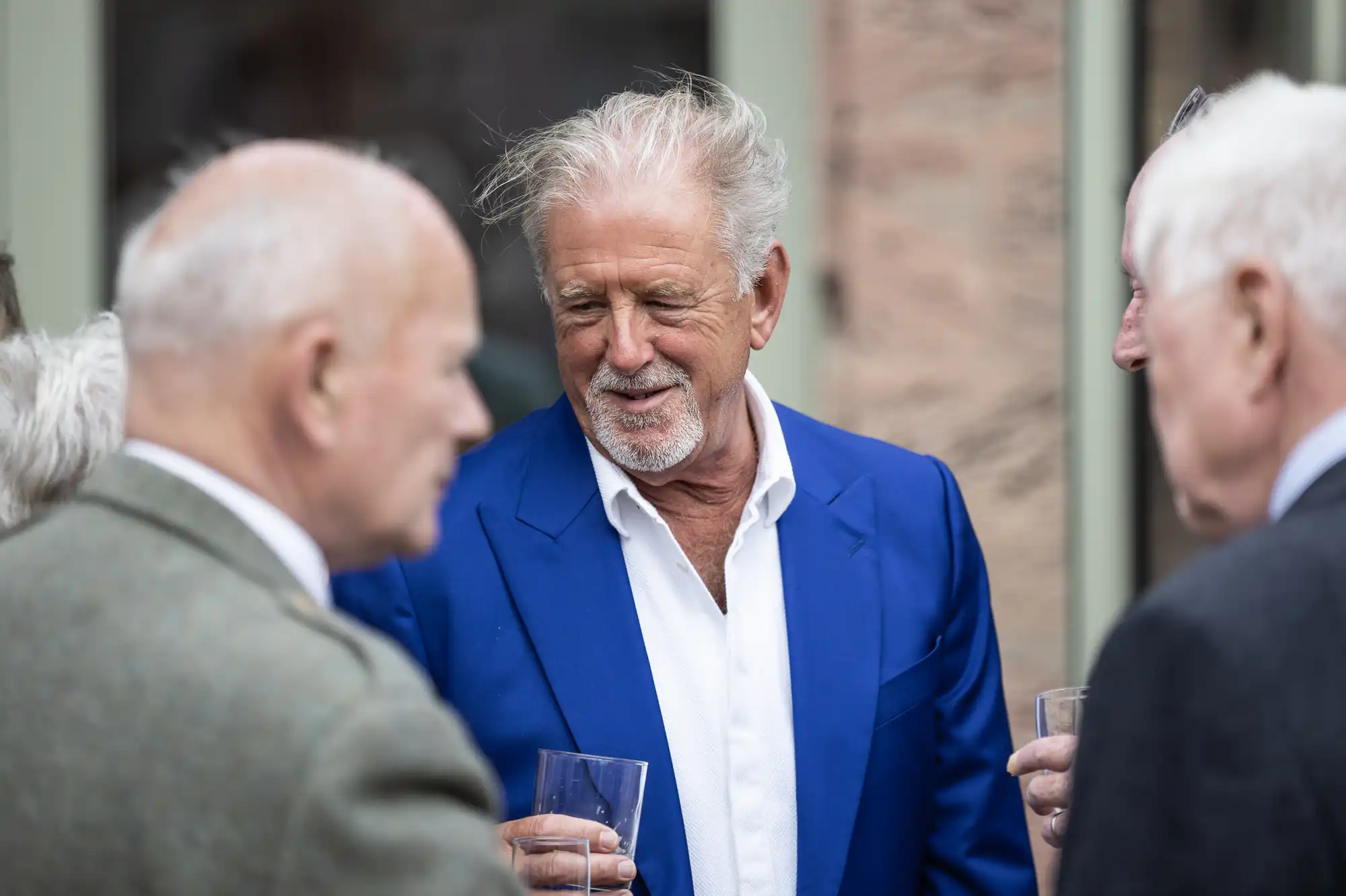 A man in a blue suit holds a drink and converses with three other men at an outdoor gathering.