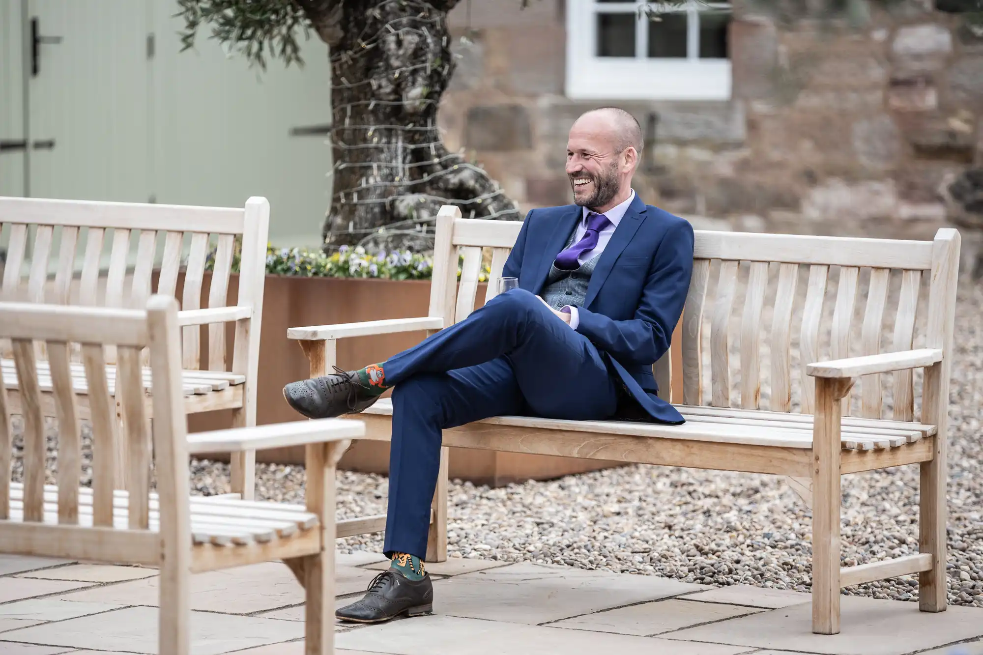 A man in a blue suit sits smiling on a wooden bench in an outdoor setting with a cobblestone ground and background.