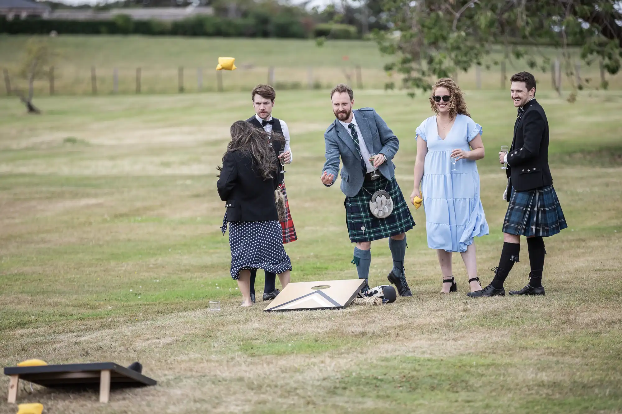 A group of people, dressed in traditional Scottish attire, are playing an outdoor game of cornhole on a grassy field. One person is in the act of tossing a bean bag towards the game board.