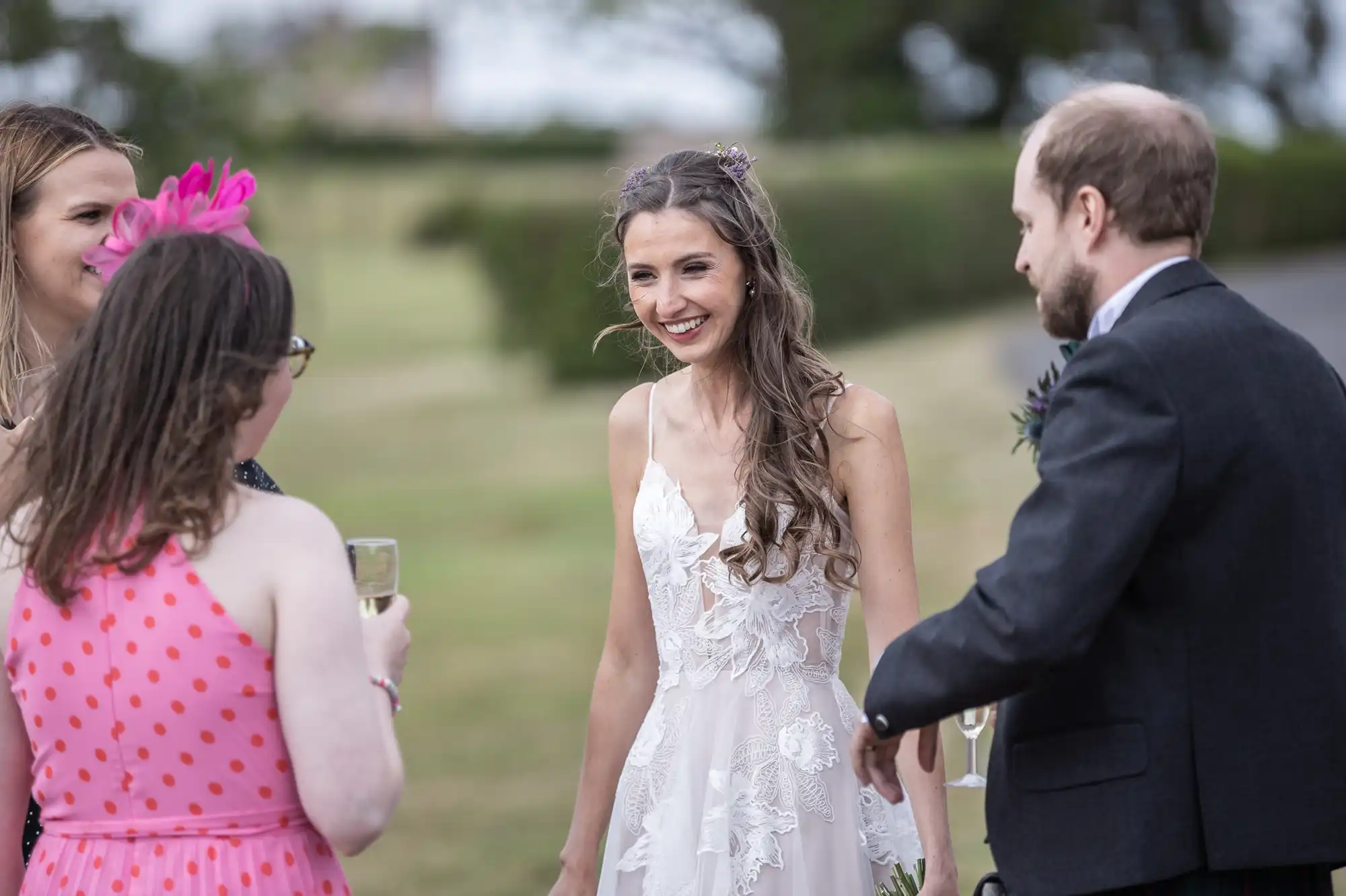 A bride in a white dress smiles and chats with three guests outdoors. The guests, two women in colorful attire and a man in a suit, hold champagne glasses.