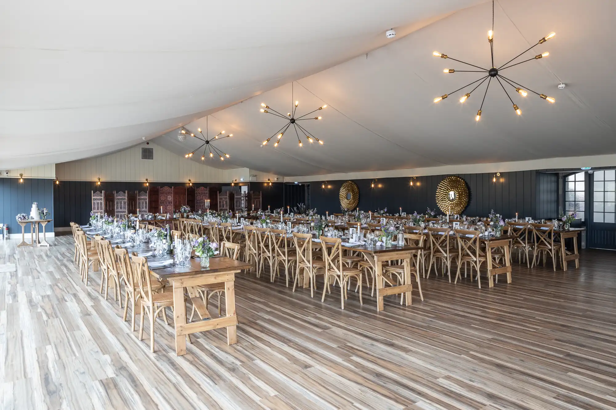 A spacious, well-lit banquet hall with wooden floors, long tables set for dining, and modern chandeliers hanging from the ceiling. Tables are adorned with floral arrangements and tableware.