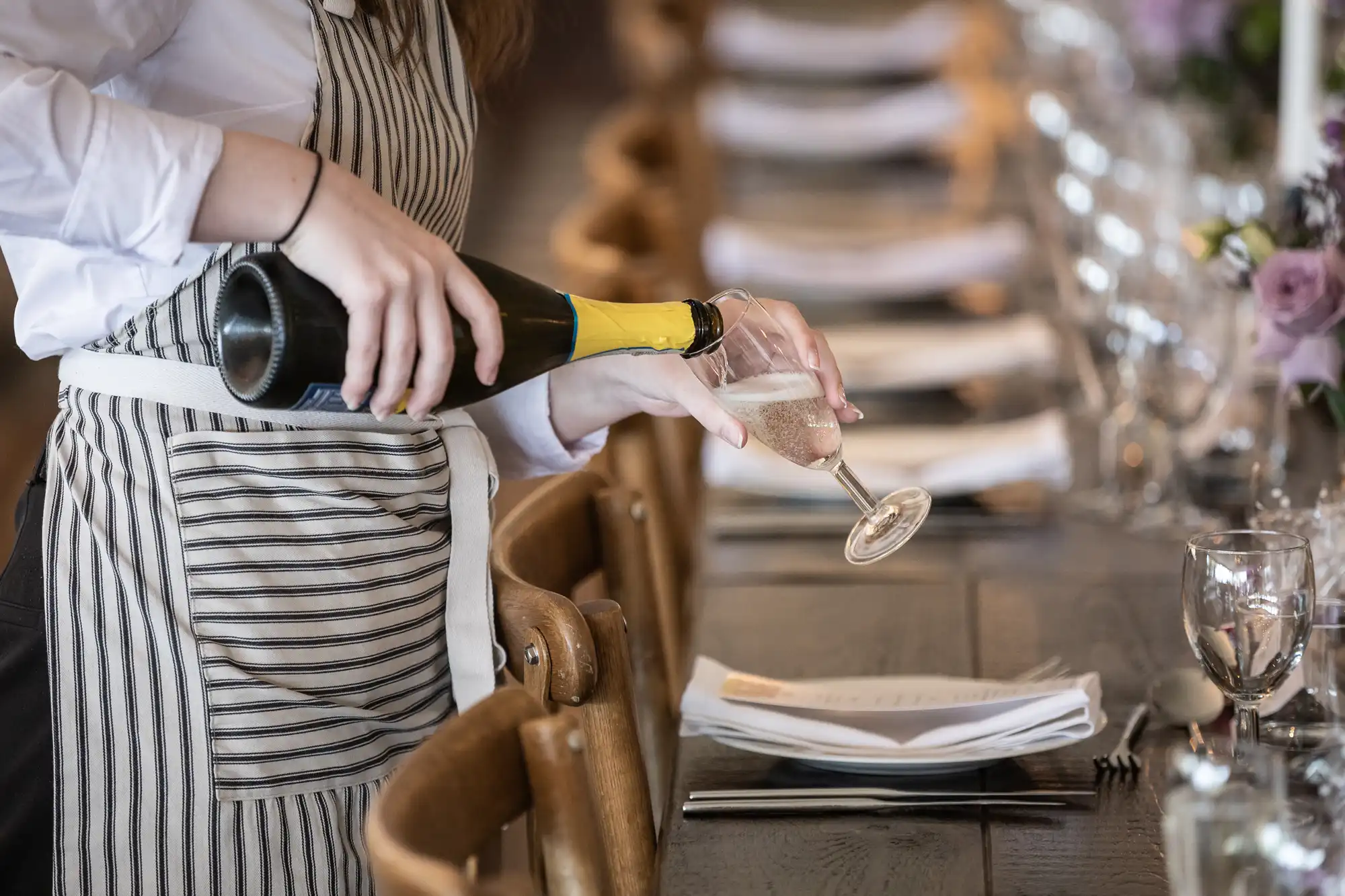 A server in a striped apron pours wine into a glass at a set dining table.