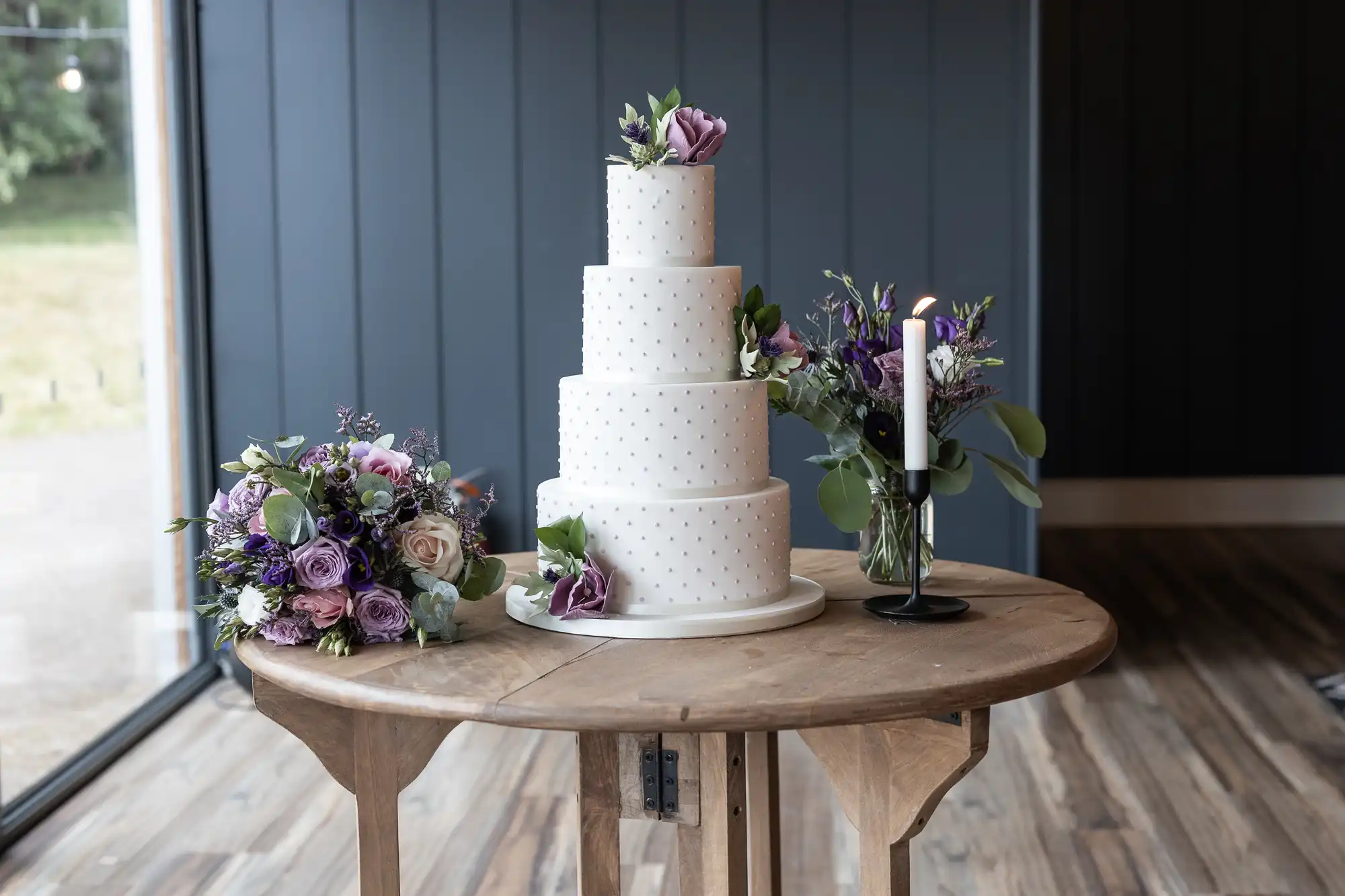 A three-tiered white wedding cake decorated with small polka dots and purple flowers sits on a round wooden table next to floral arrangements and a single purple candle.