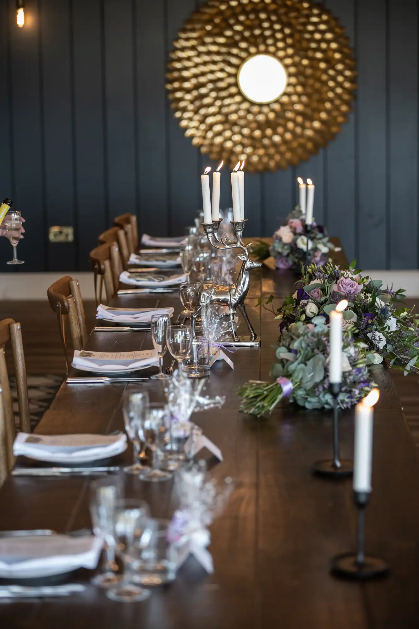 A long dining table set with plates, glasses, floral arrangements, and lit candles in holders against a dark blue paneled wall with a large decorative mirror.