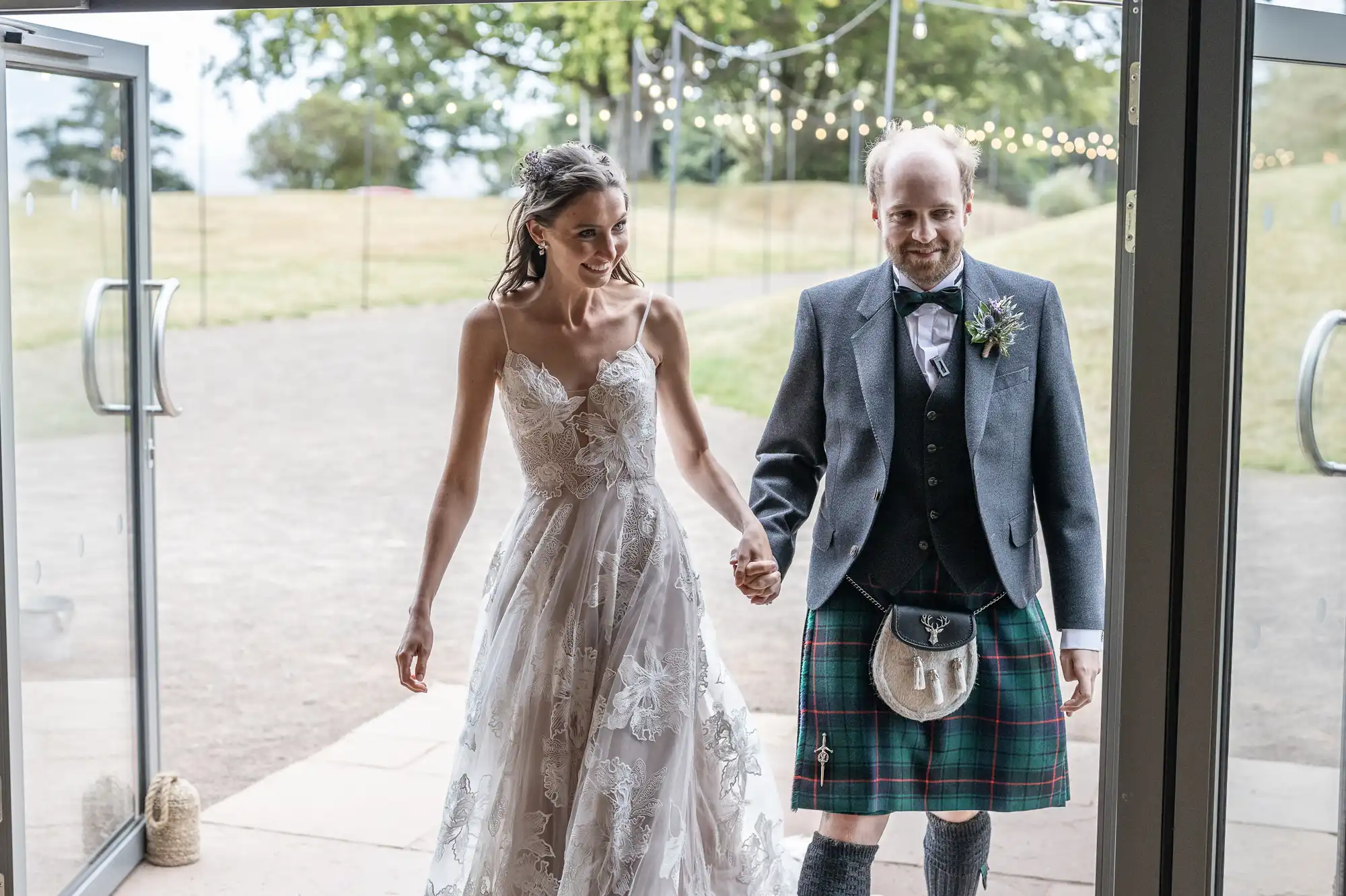 A groom in a kilt and a bride in a white dress hold hands as they enter a venue through glass doors. String lights are visible in the background.