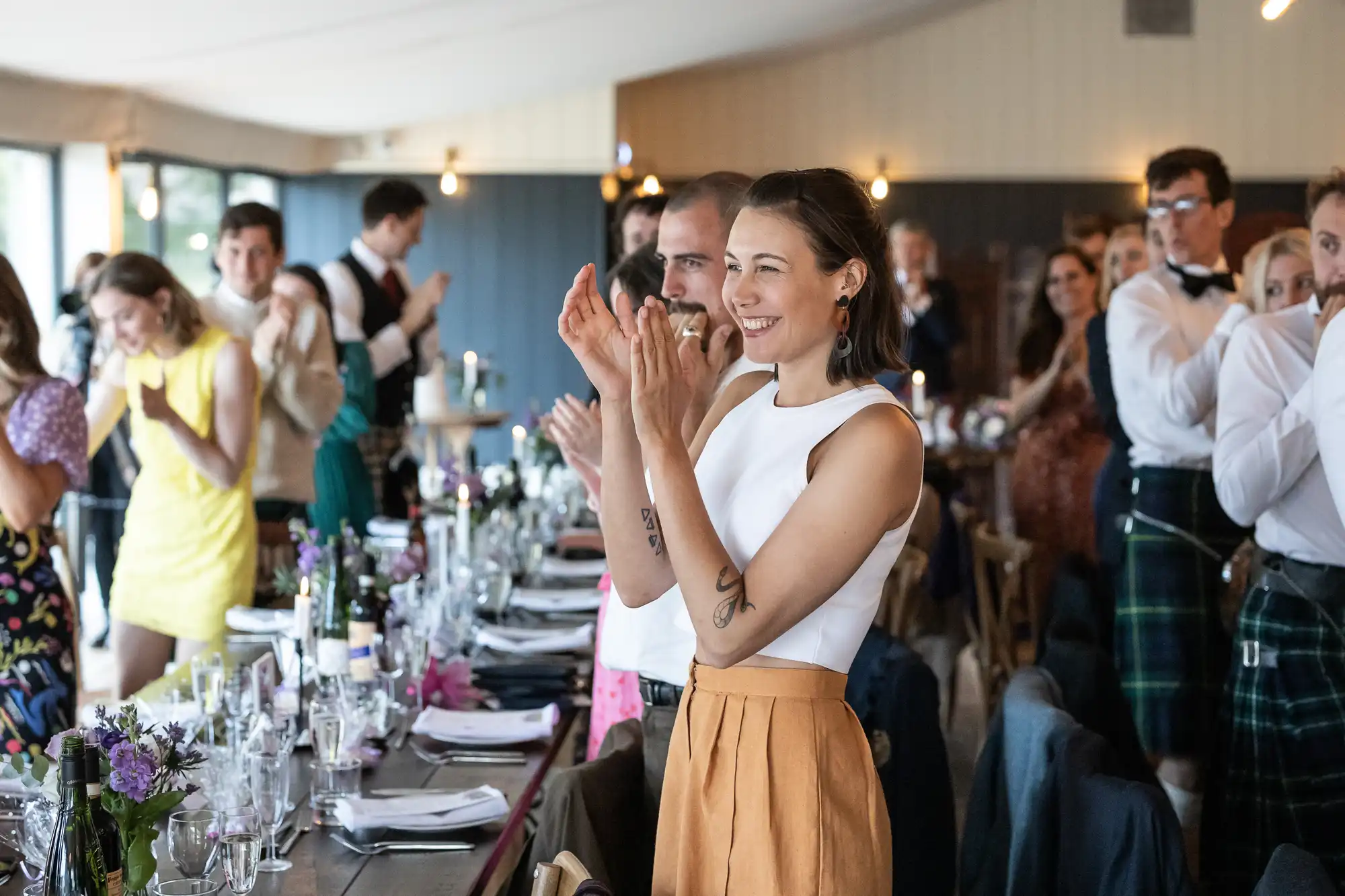 People standing and clapping in a well-lit room with a long dining table set for a meal. Some individuals are wearing formal attire.