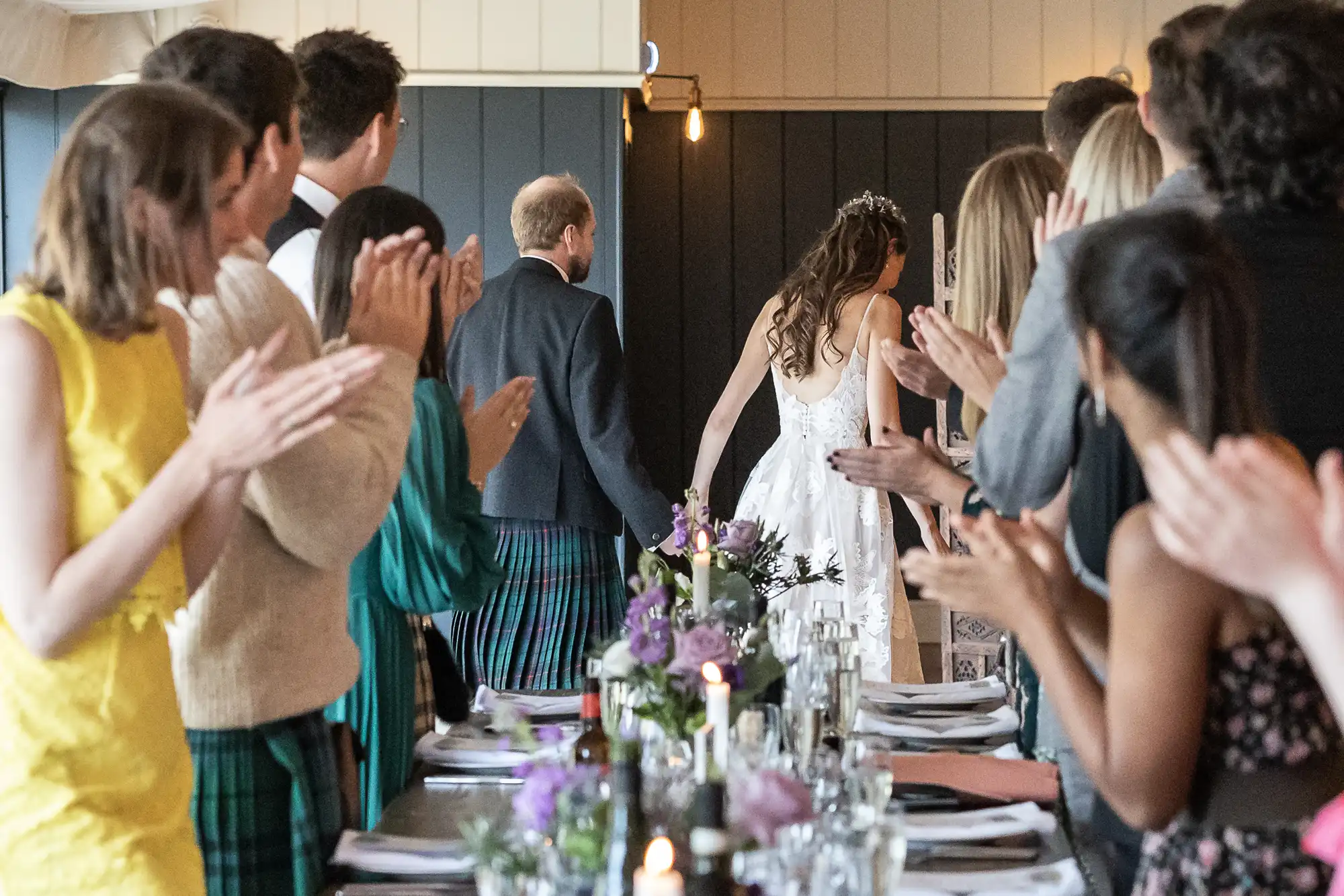 A newlywed couple walks hand-in-hand down an aisle while guests on both sides stand and applaud. The bride is in a white dress, and the groom is in a suit with a kilt.