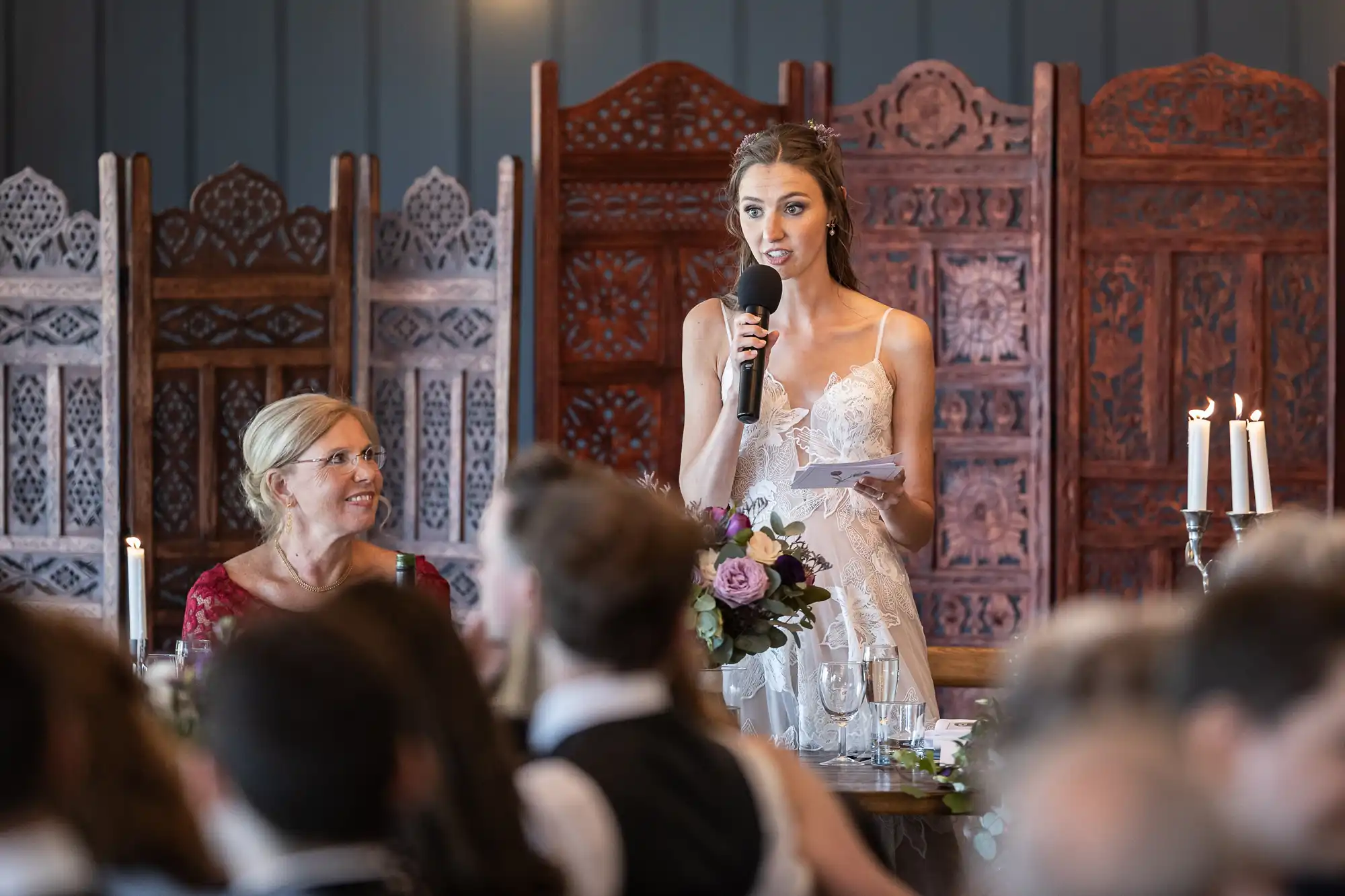 A woman in a white dress speaks into a microphone while holding notes, standing in a room with wooden partitions. Another woman in a red dress sits at a table, looking up at the speaker.