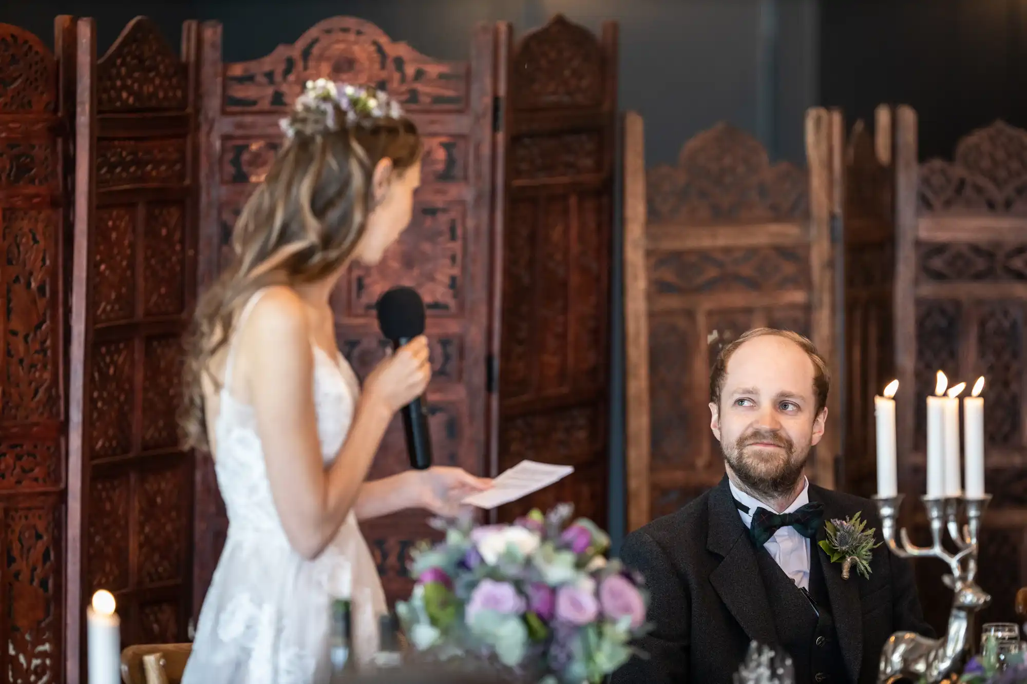 A woman in a white dress is giving a speech with a microphone while a man in a suit sits and listens. They are in a room with ornate wooden dividers and a table with flowers and candles.