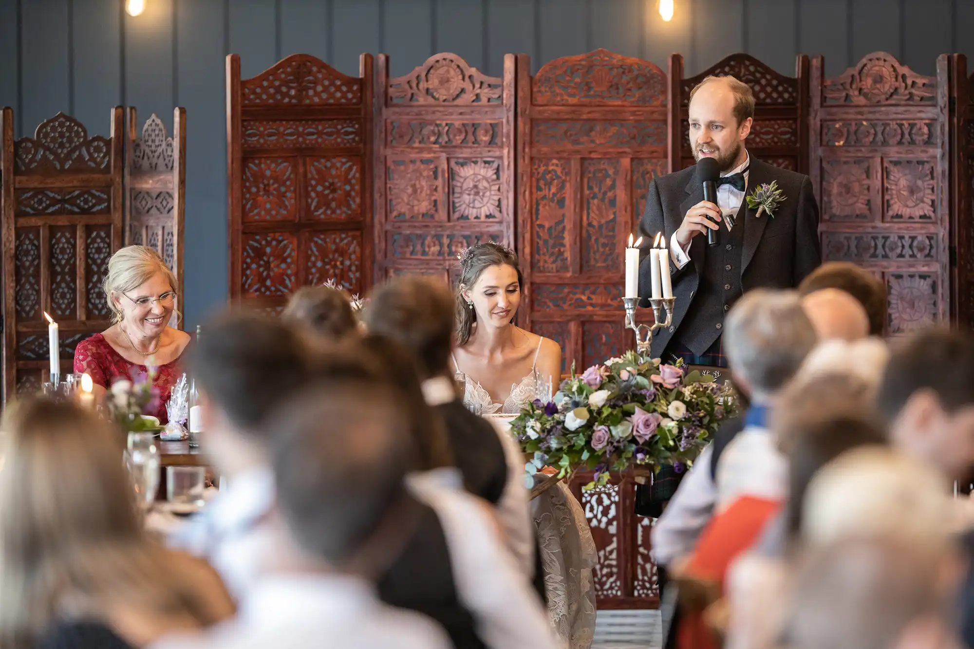 A man speaks into a microphone next to a bride, who is sitting and smiling. Another woman is seated beside the bride. The scene appears to be a wedding reception with guests seated and listening.