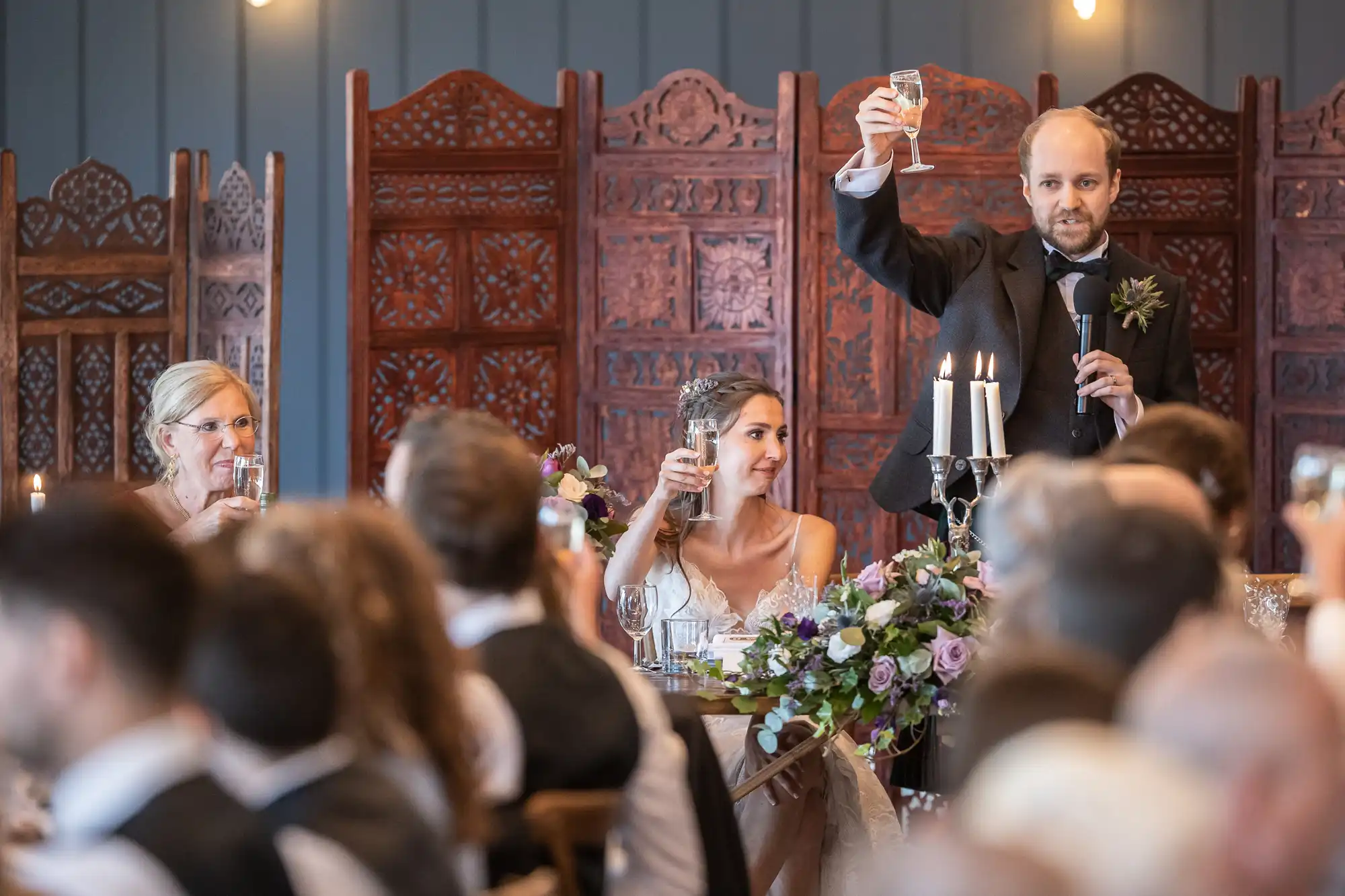 A person in a suit stands and raises a glass for a toast at a formal event, while others seated around a table also lift their glasses. Decorative wooden panels are in the background.