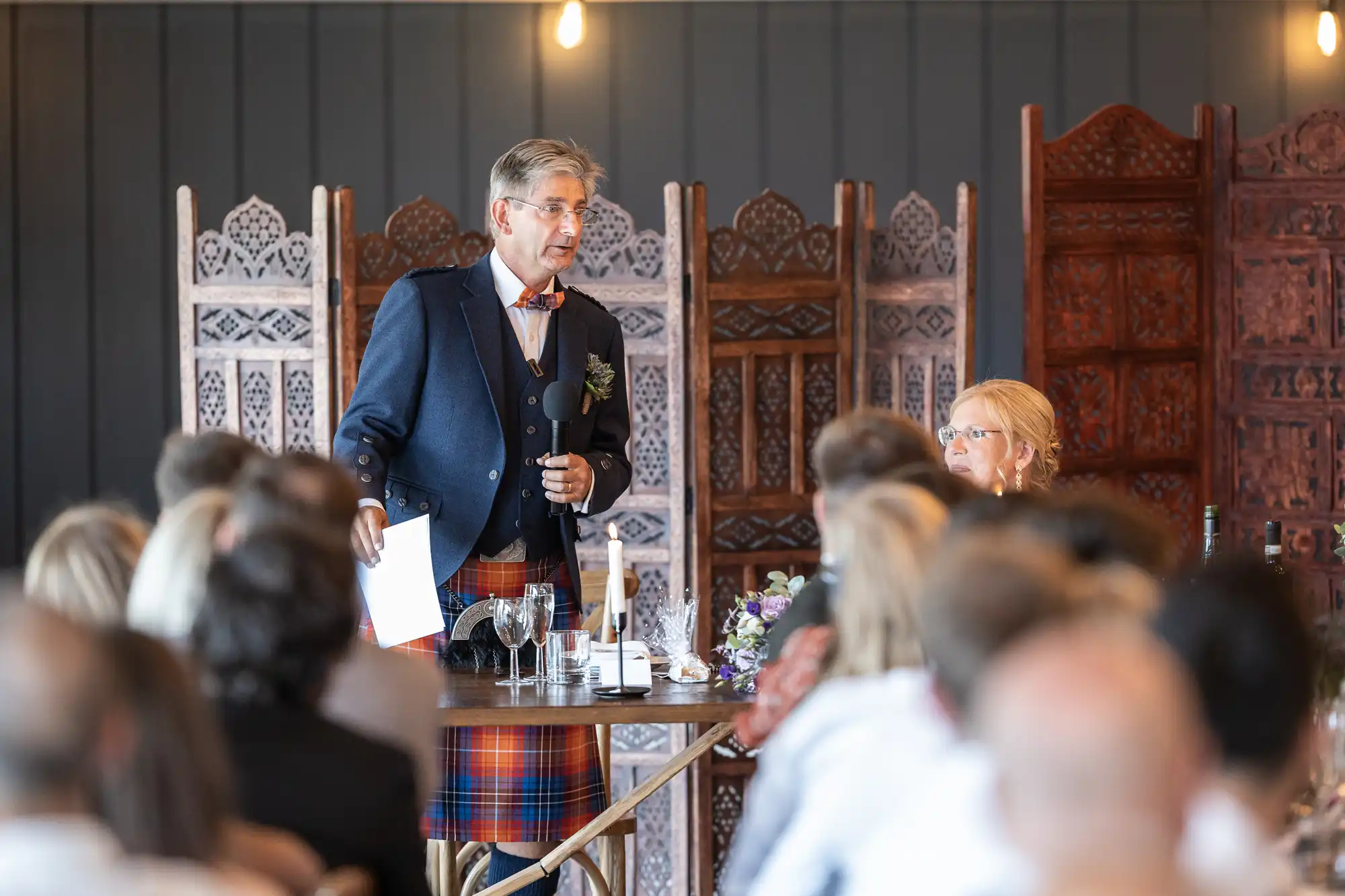 A man wearing a suit jacket and tartan trousers speaks into a microphone at a formal gathering, while an audience listens. Decorative room dividers are in the background.