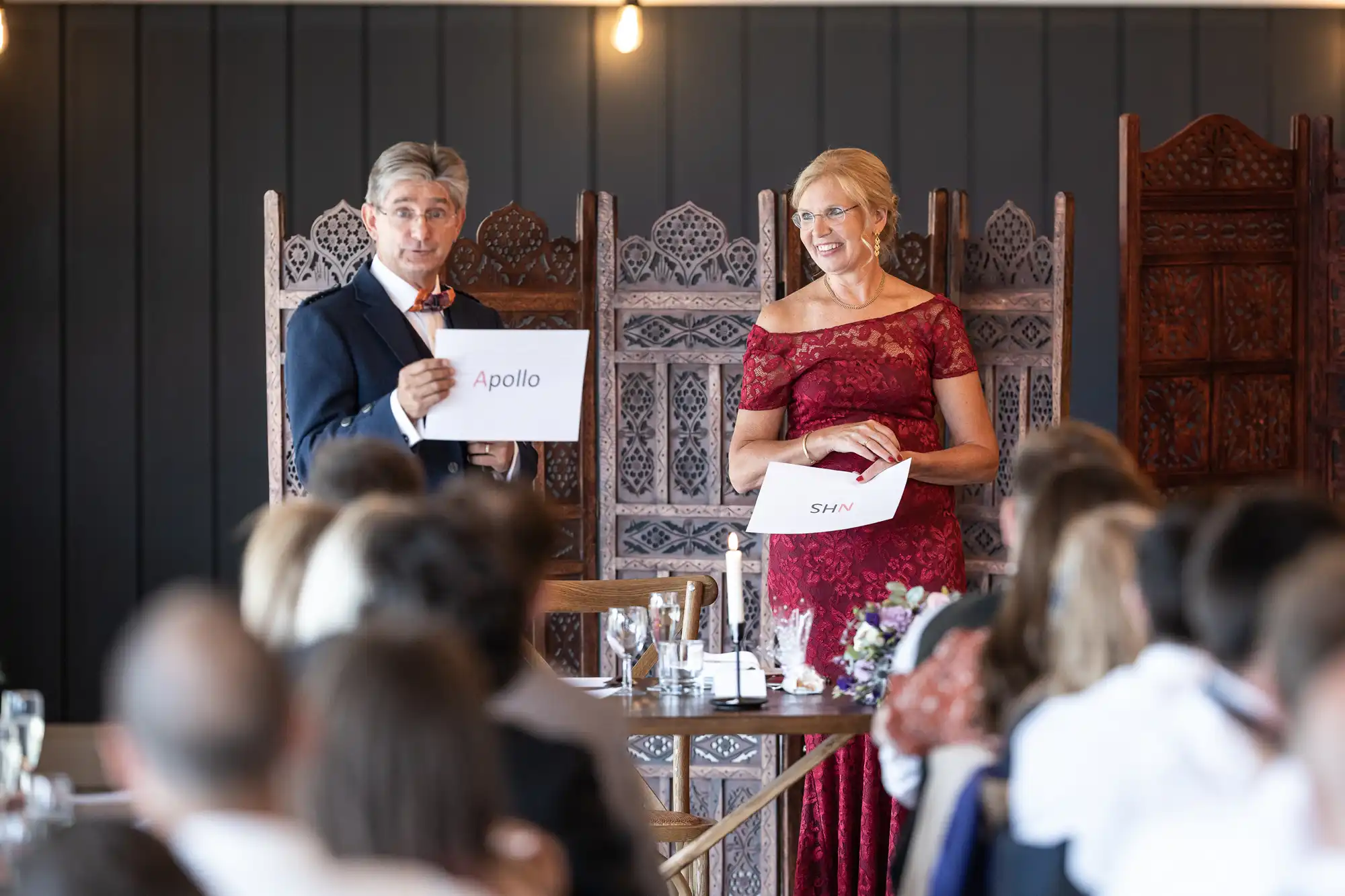 Two people stand at the front of a room holding signs that read "Apollo" and "SHN." The man is in a suit and the woman in a red dress. An audience is seated in front of them.