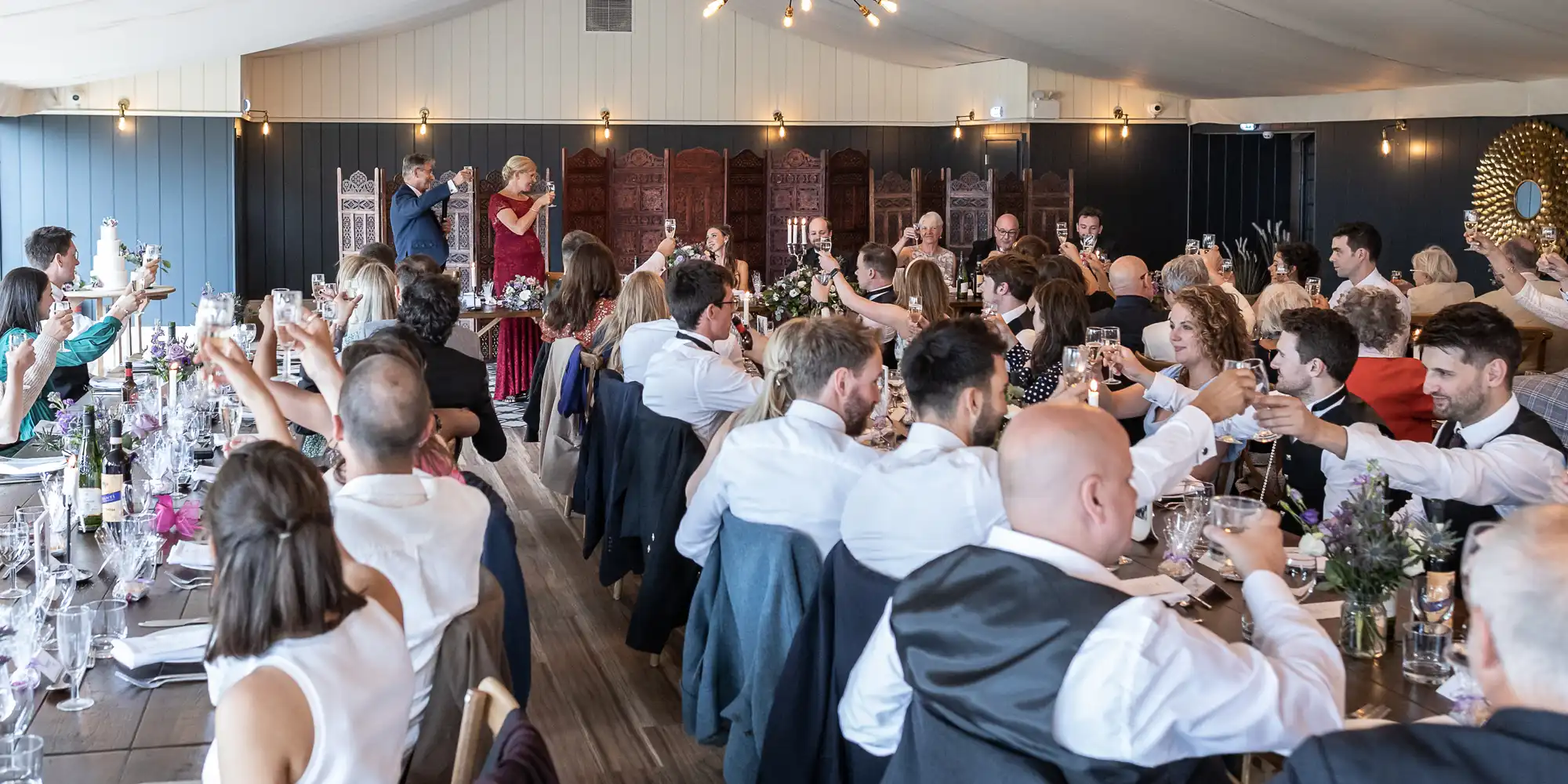 A large group of seated people at a formal event indoors, raising glasses in a toast, with two speakers behind a lectern at the front of the room.