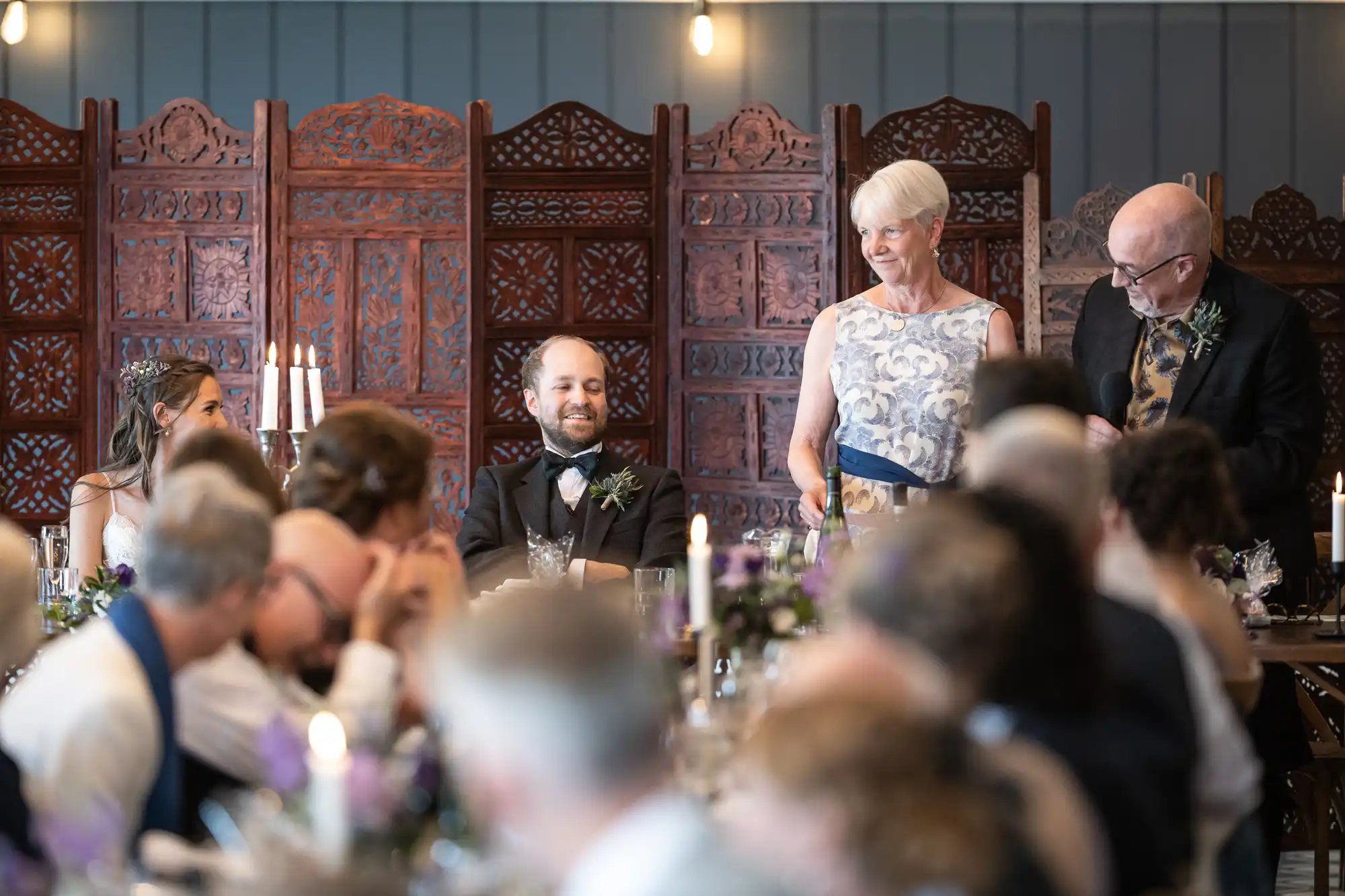 A wedding reception with seated guests listening to a speech. A couple stands to the right of the room, while the bride and groom are seated at the head table.