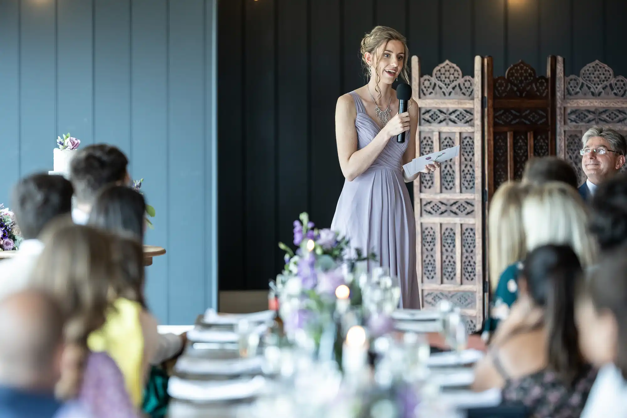 A woman in a light purple dress stands and speaks into a microphone at a formal event, with seated guests and decorated tables in the foreground.