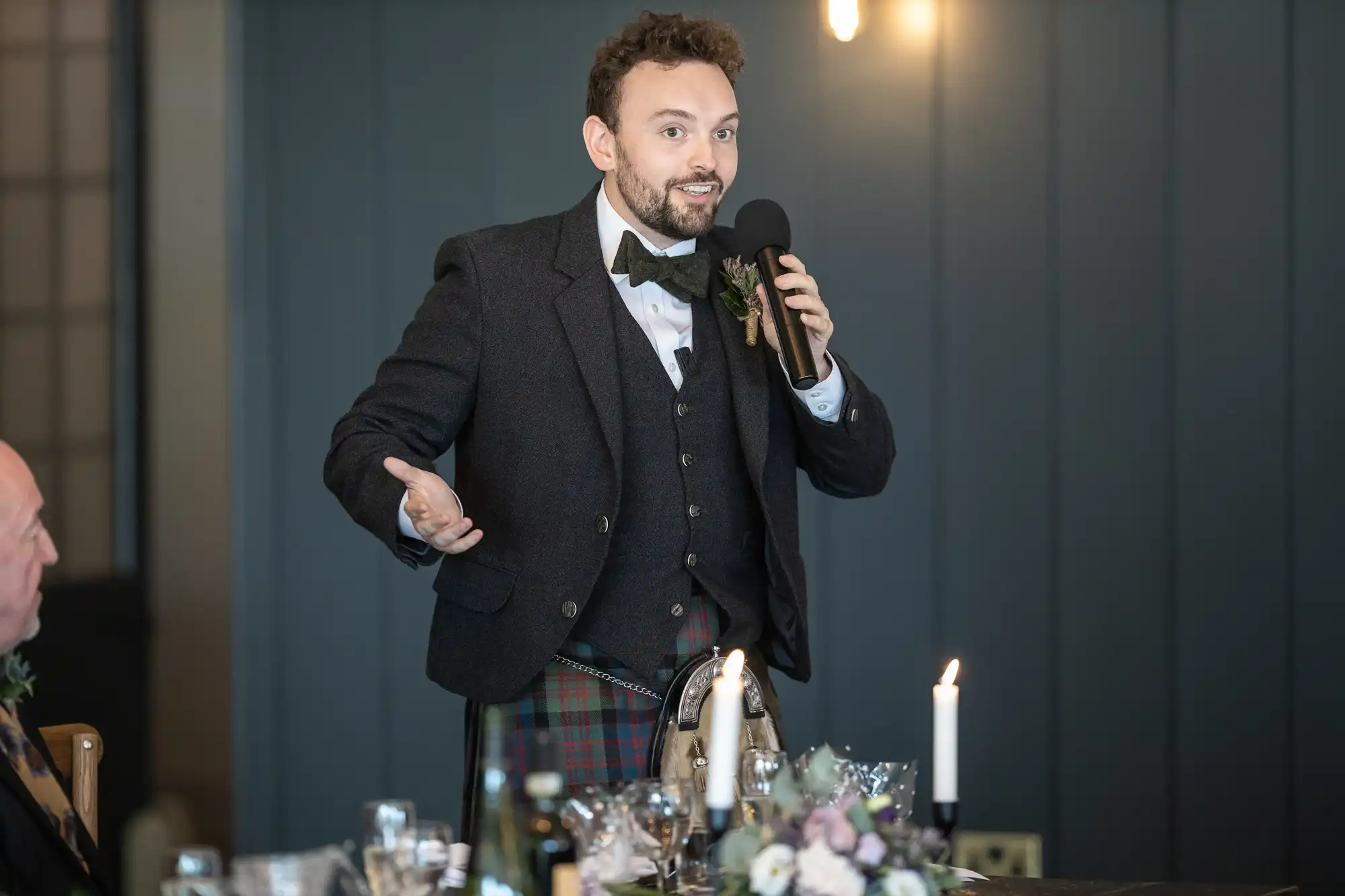 A man in formal attire speaks into a microphone while standing at a table decorated with candles and floral arrangements.