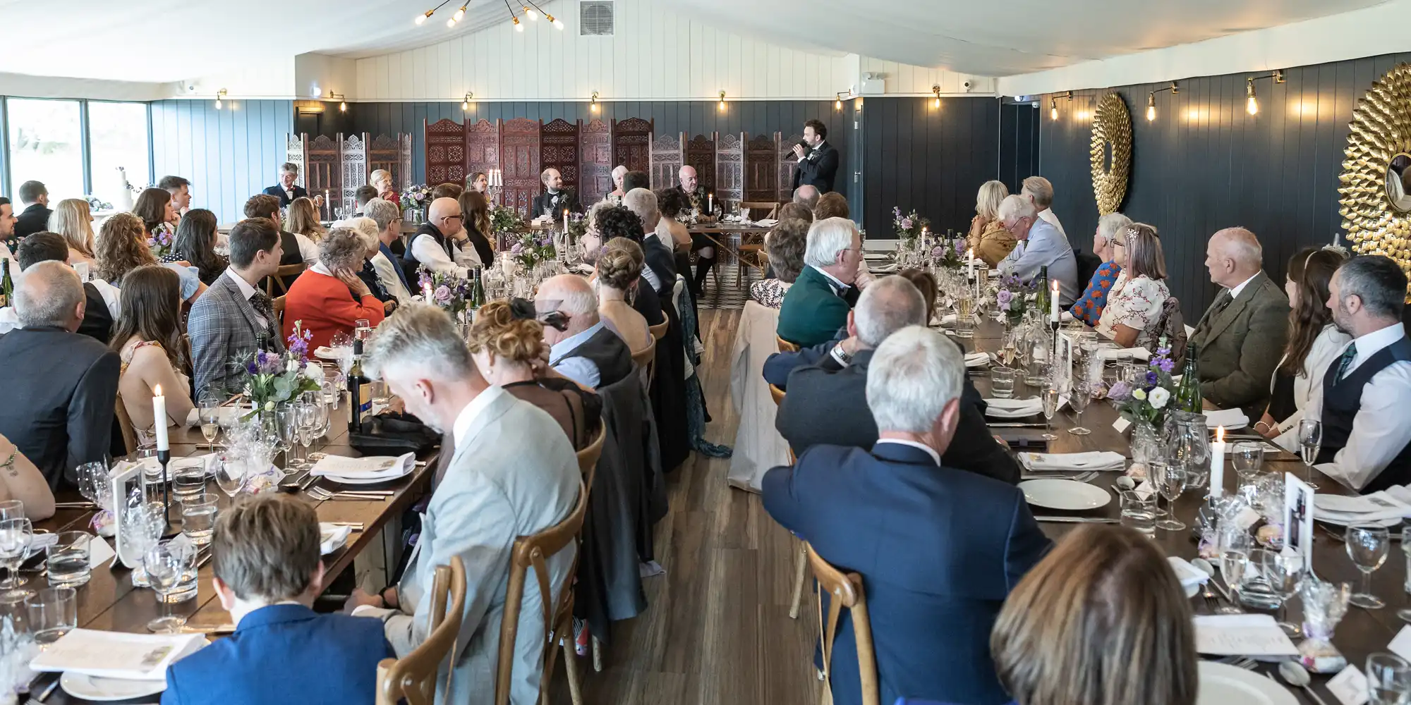 A large group of people seated at tables in an indoor venue, attentively listening to a speaker at the front of the room. The tables are decorated with floral arrangements and place settings.