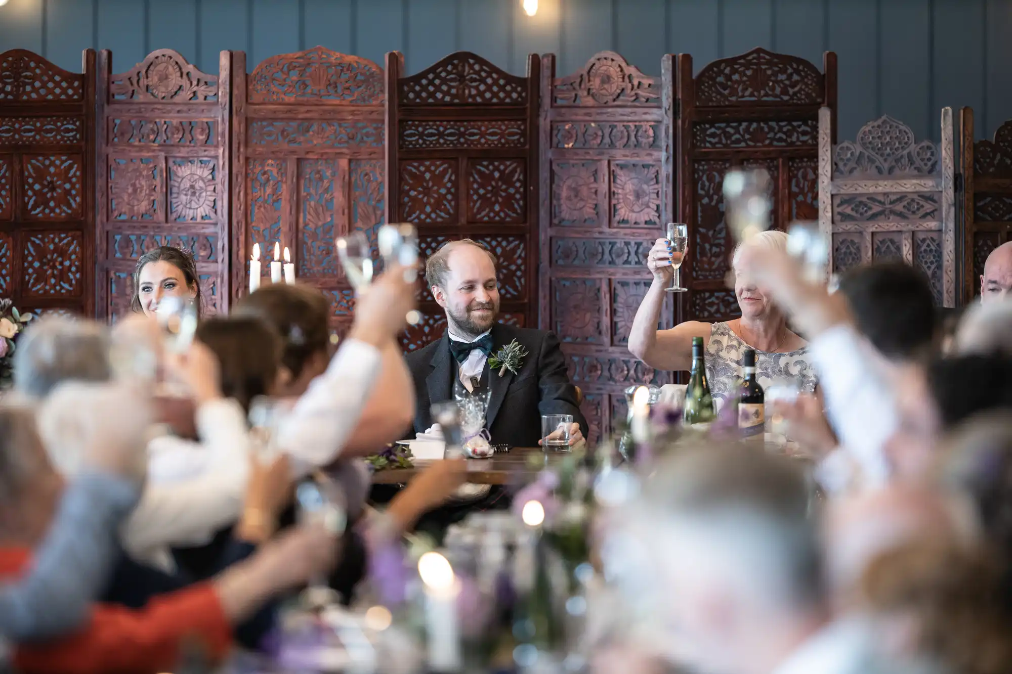 A man in a suit sits at a table with people around him raising glasses for a toast in a decorated indoor setting.