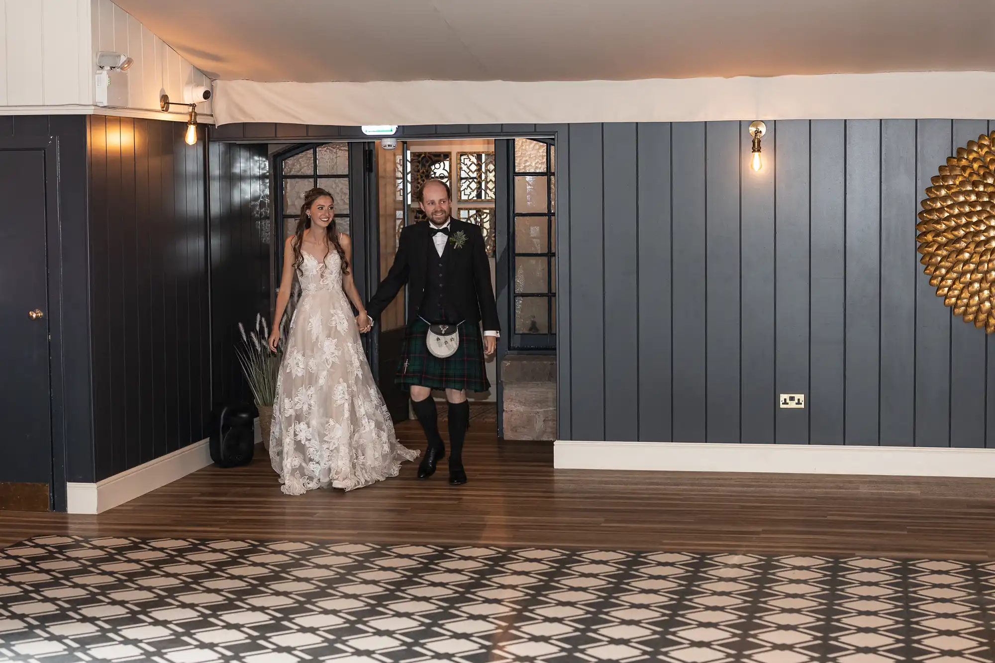 A bride in a white dress holding hands with a groom in a kilt walk into a room with dark blue walls and patterned flooring.