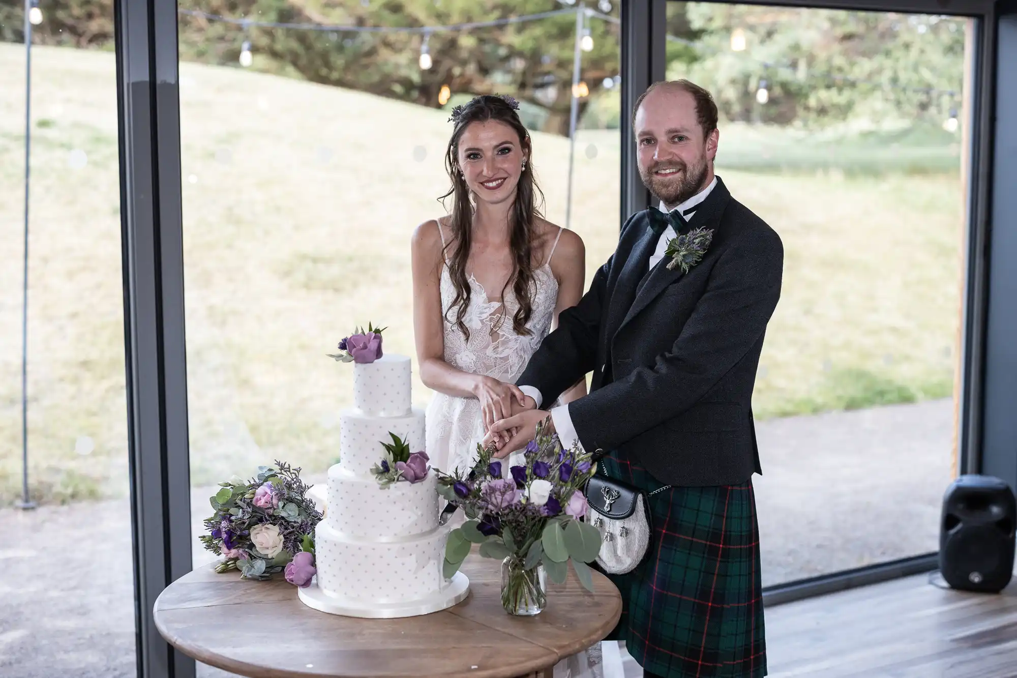 A bride and groom stand together, cutting a white three-tiered wedding cake decorated with purple flowers. The groom is dressed in a traditional kilt, and they both smile at the camera.