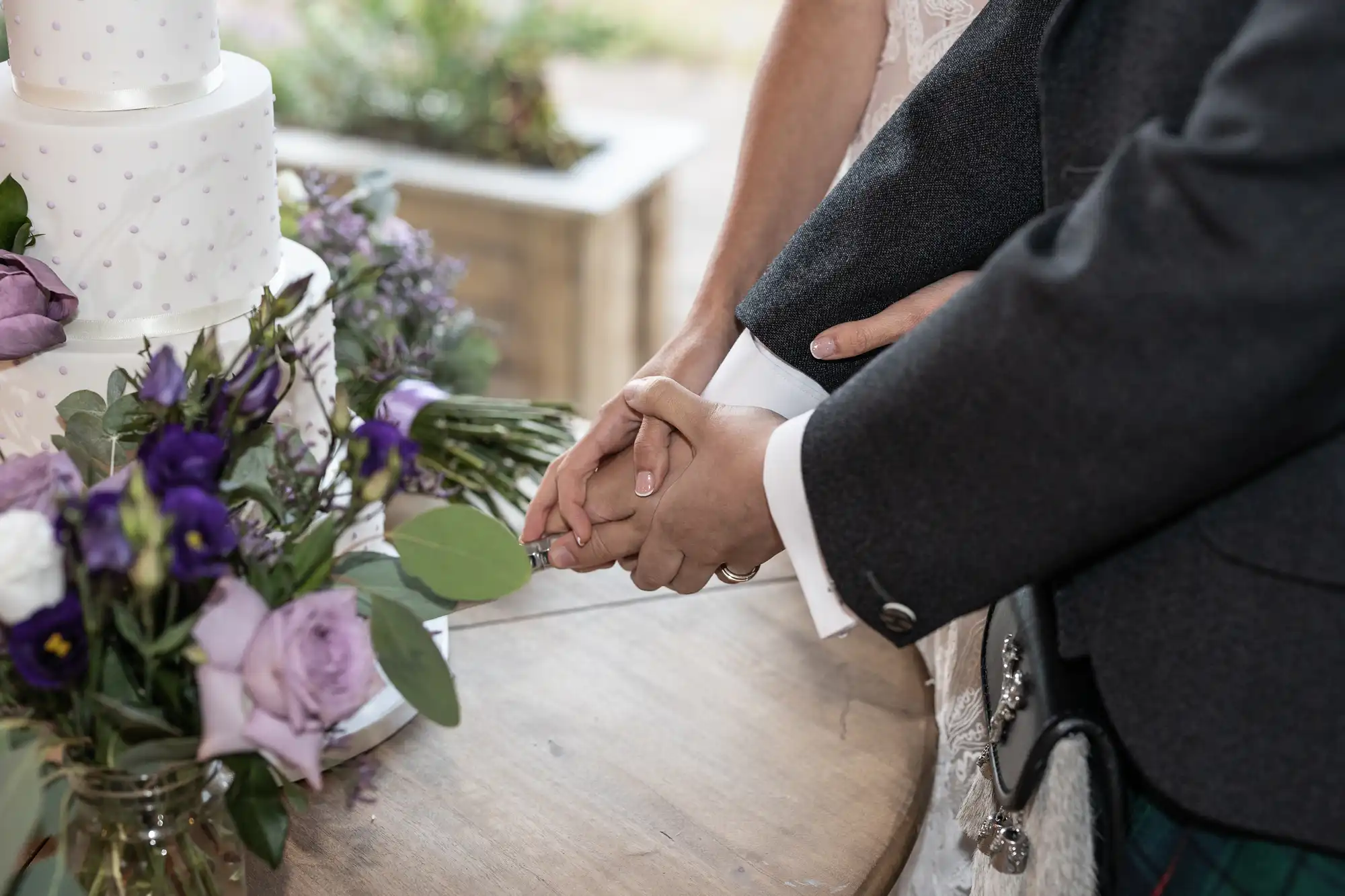 Two people, dressed in formal attire, hold hands next to a table with a floral arrangement and a white, multi-tiered cake.