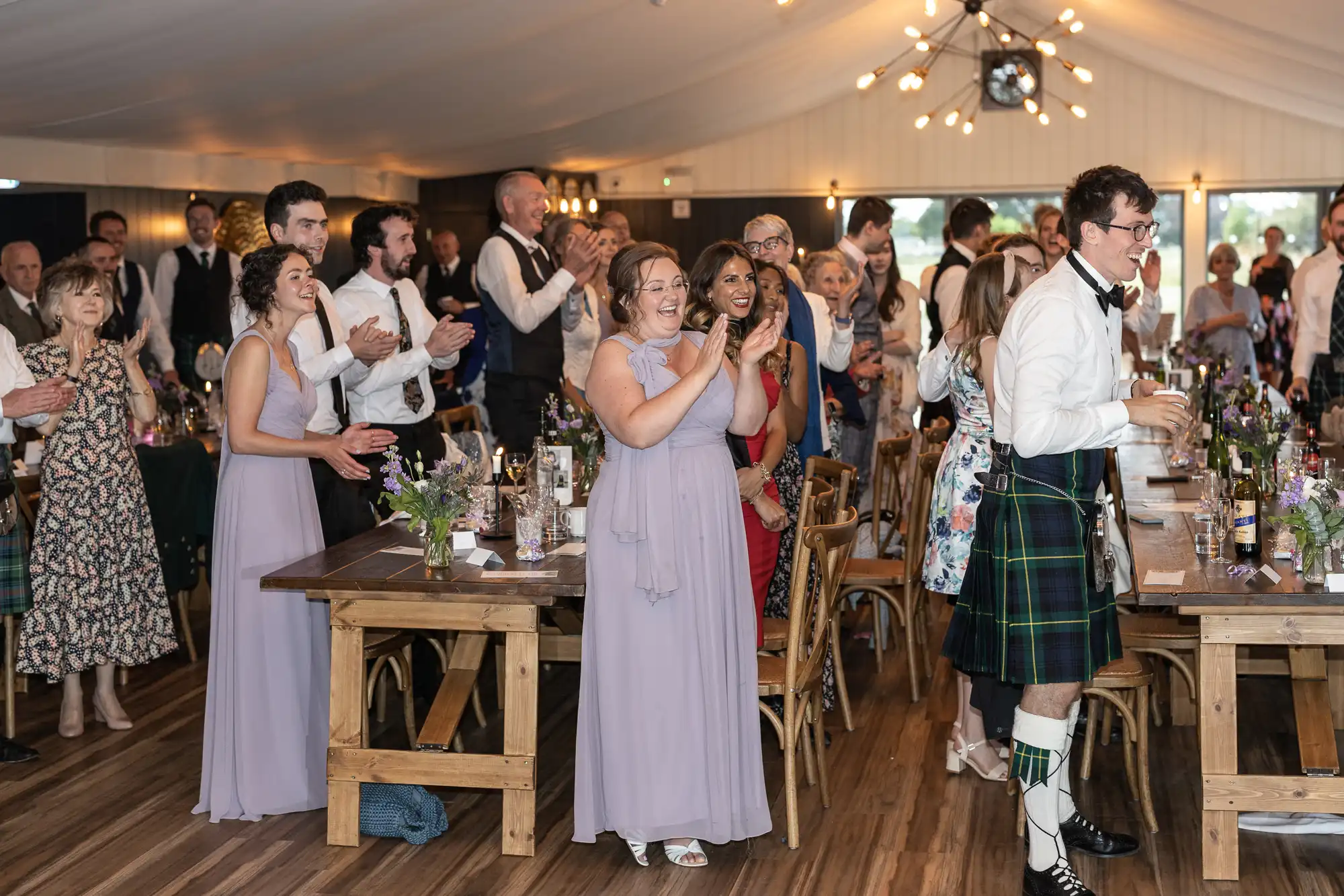 A wedding reception with guests standing and clapping around wooden tables, some wearing formal attire and kilts, in a warmly lit venue.