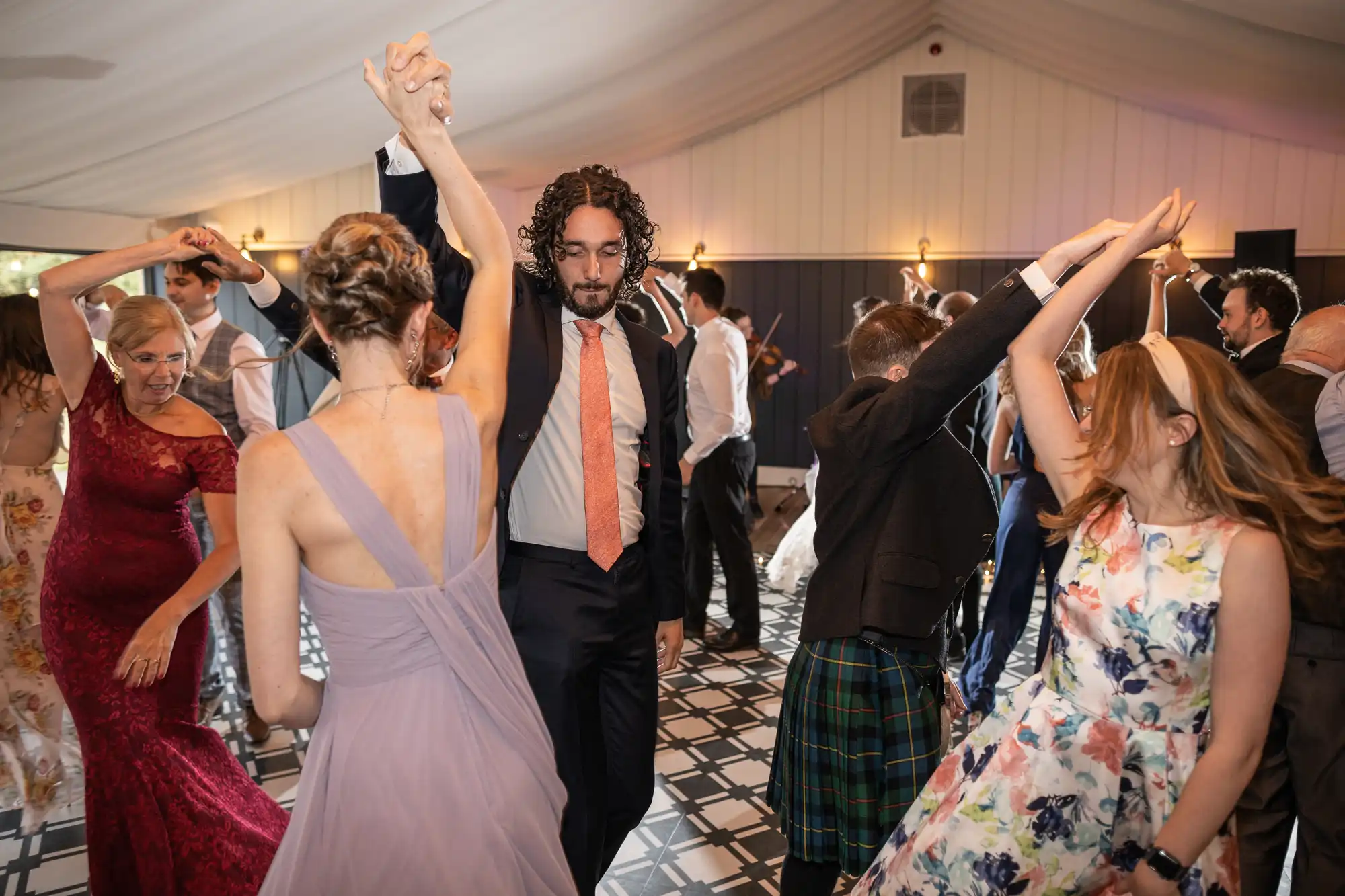 A group of people dressed in formal attire are dancing under a tent with a checkered floor. Many of them have their arms raised, forming arches.