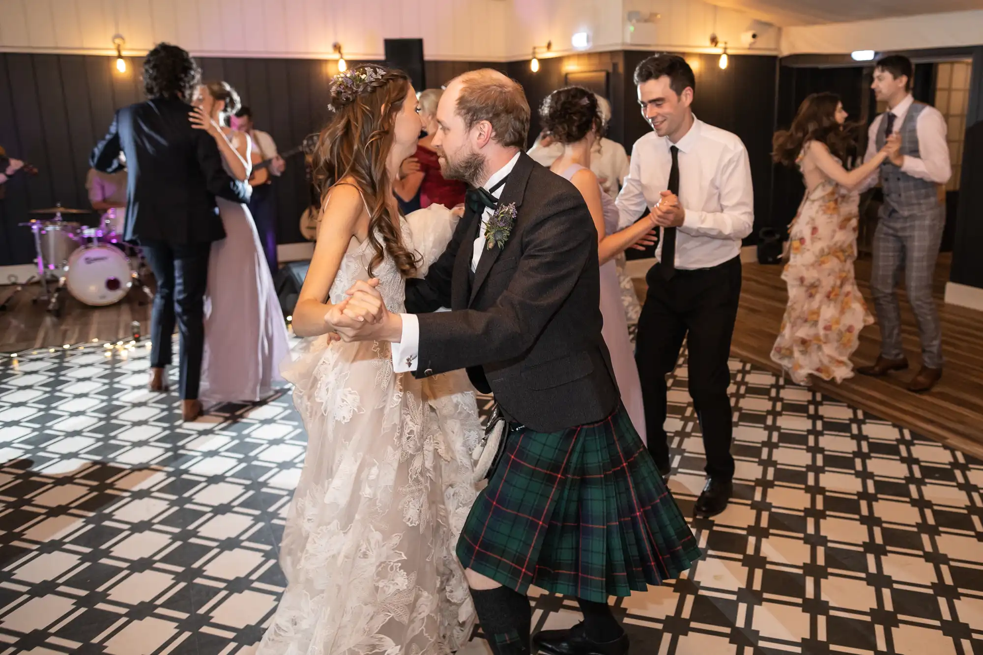 People dancing at an indoor event. A couple dances in the foreground, one in a white gown and the other in a plaid kilt. Other attendees are dancing and conversing in the background.