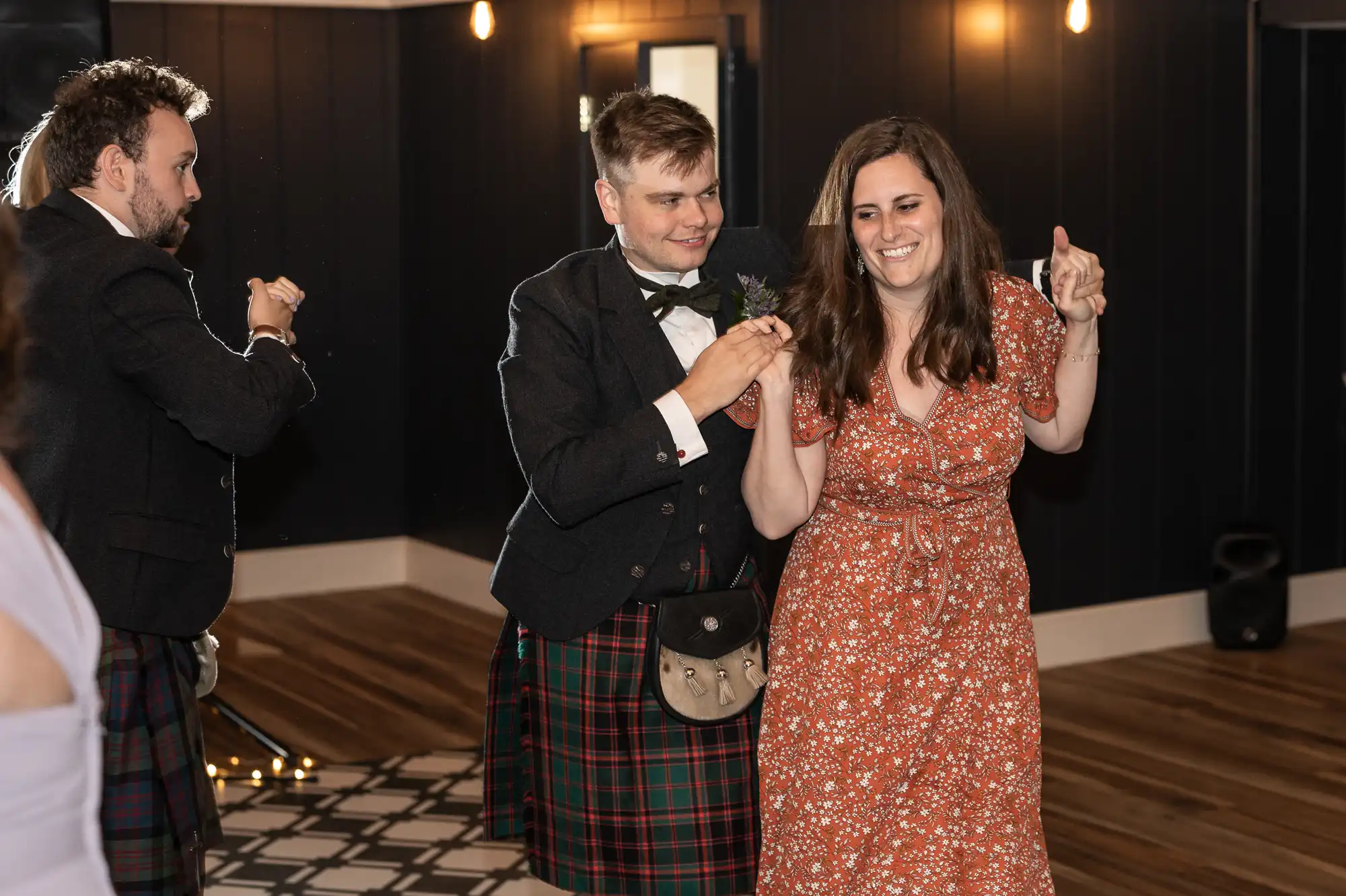 Two men in kilts and a woman in a red dress dance together in a warmly lit room with wooden floors and dark walls.