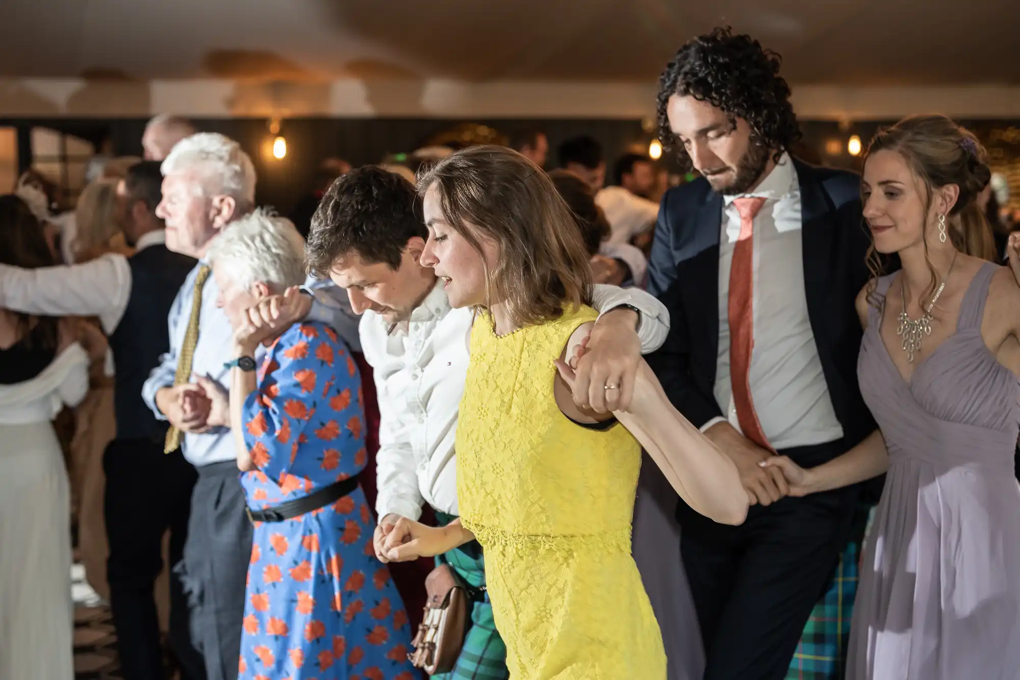 A group of people, dressed in formal and semi-formal attire, are dancing closely together in a well-lit indoor setting.