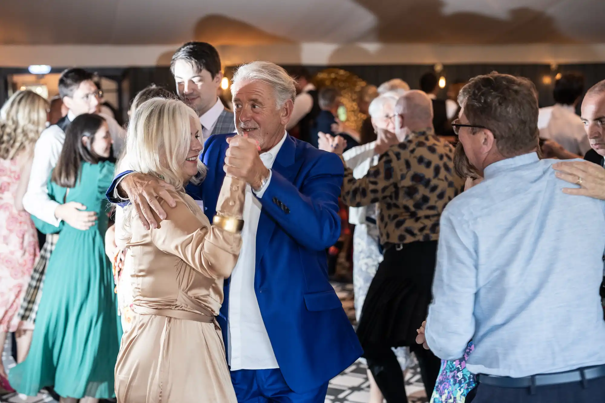 A group of people are dancing at an indoor event. In the foreground, an older couple is dancing closely, with the man in a blue suit and the woman in a beige dress. Other guests are dancing in the background.