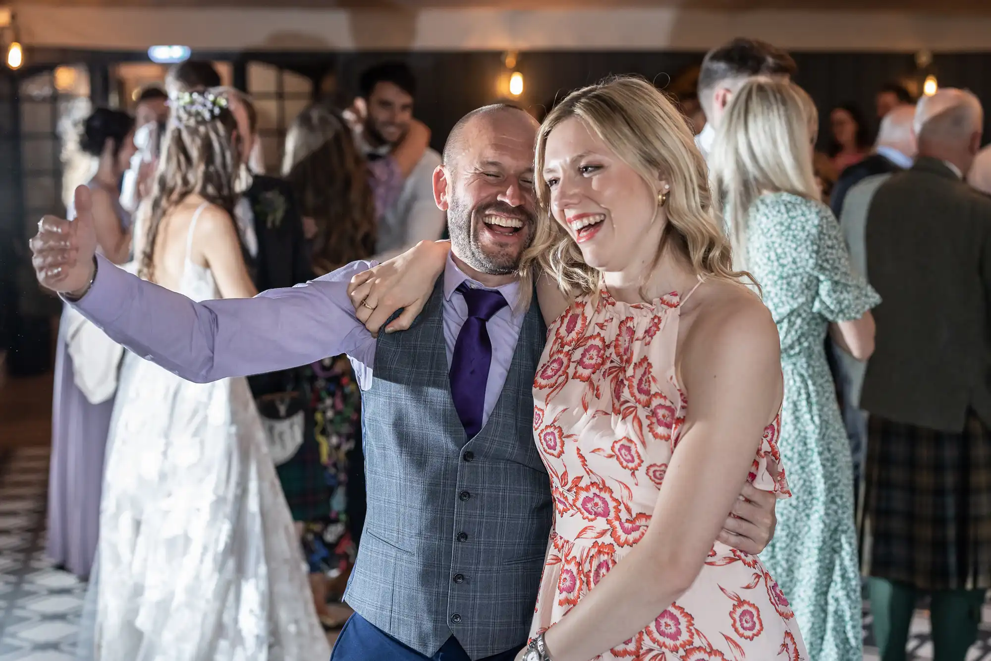 A smiling couple poses together with their arms around each other at a lively indoor event. People in the background are engaged in conversation and activities.