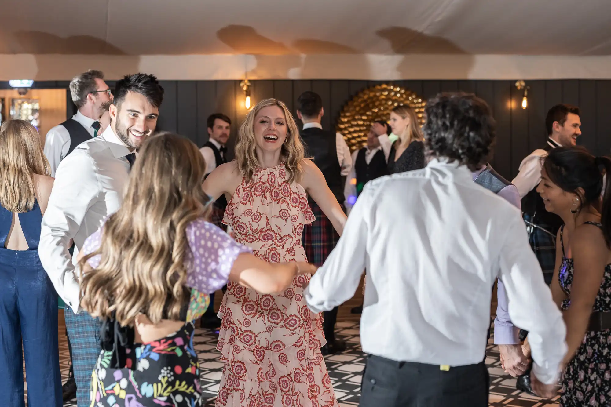 A group of people are dancing and holding hands at an indoor event. They appear to be enjoying themselves, with smiles and laughter. The room is decorated with a patterned floor and a wall ornament.