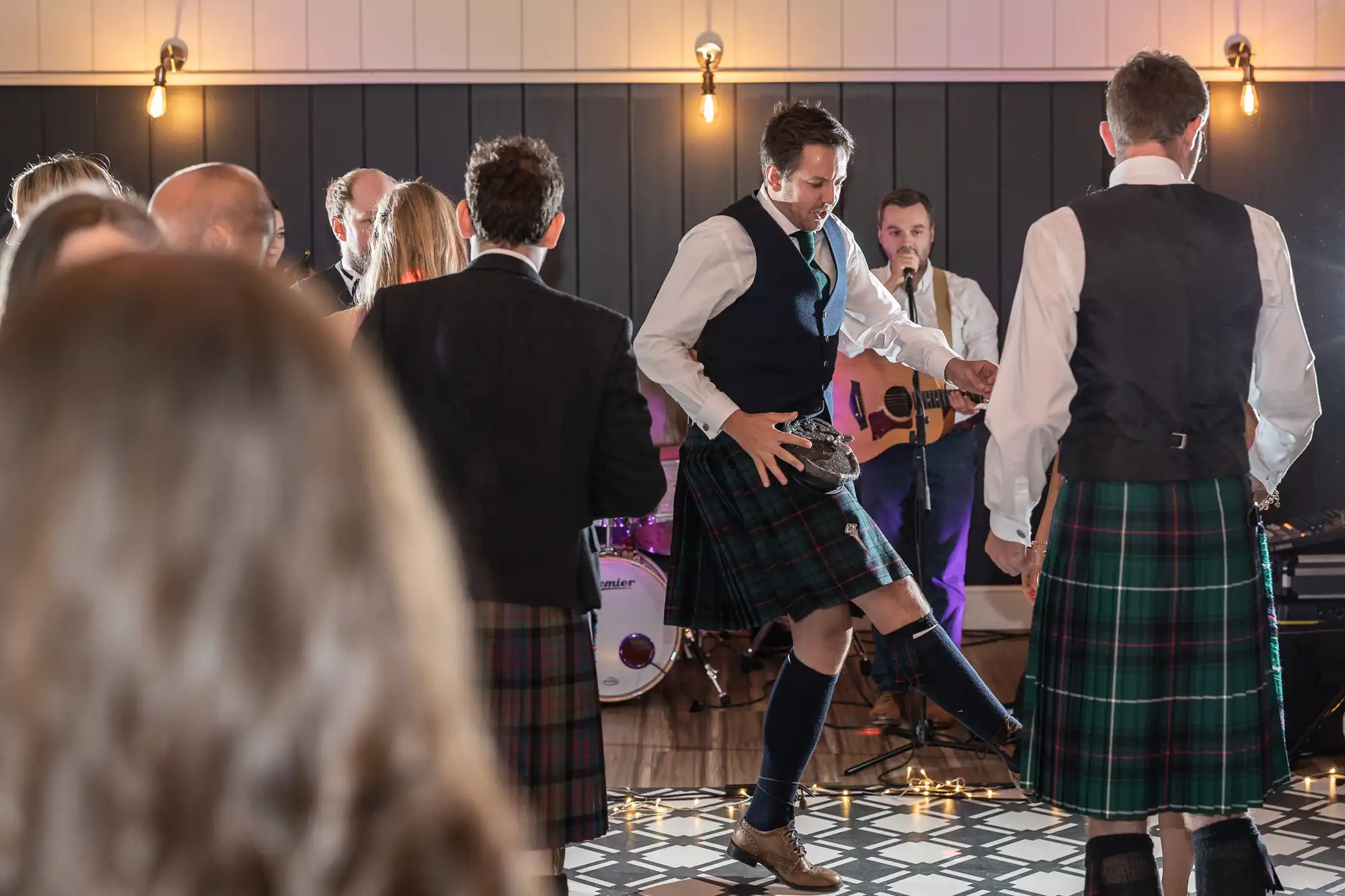 A group of people wearing kilts dance and play instruments at a gathering, with a band performing in the background.